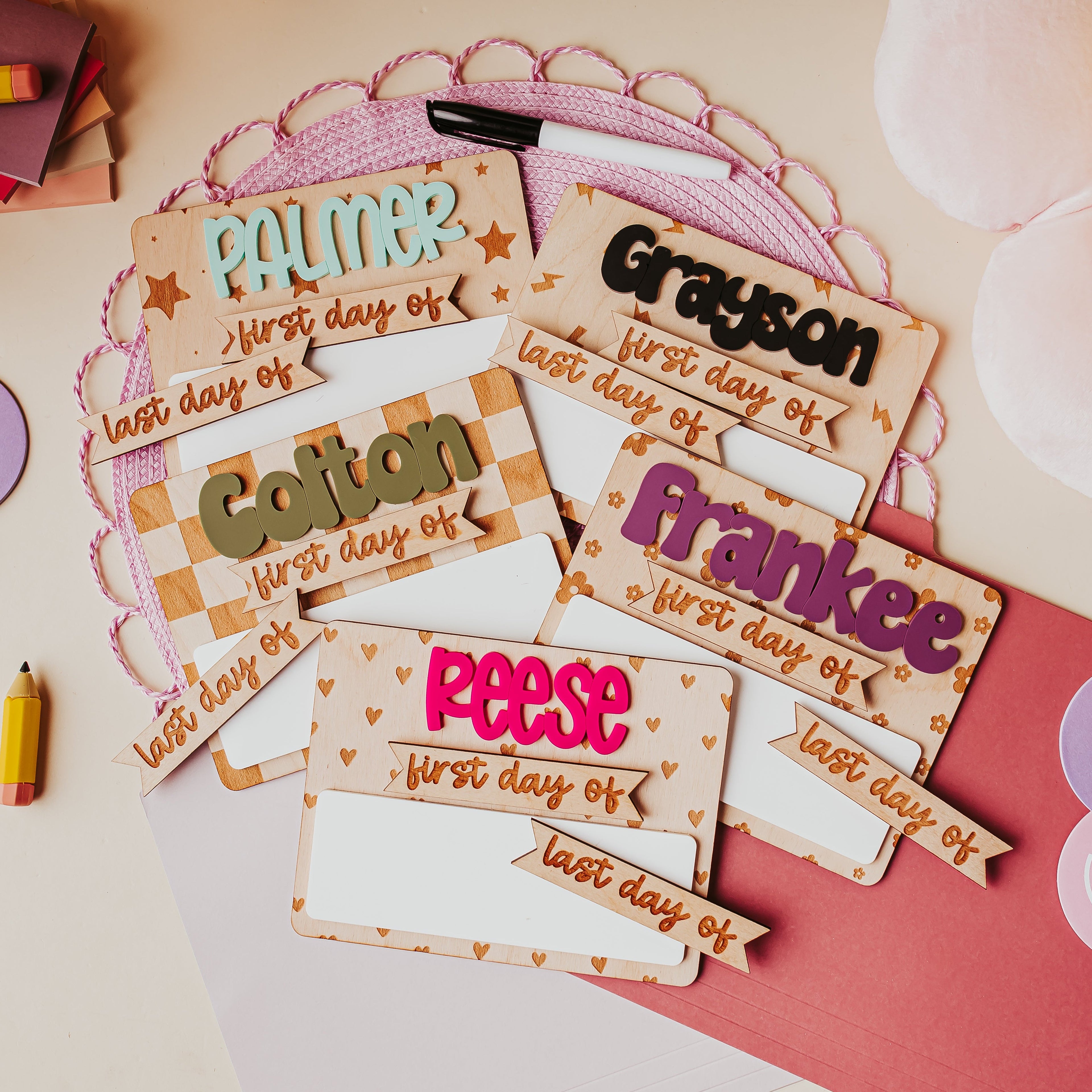 A styled flat-lay of personalized first-day-of-school dry erase signs on a light beige surface with a pink circular doily, a dry erase marker, a pink folder , and pastel smiley face post-its.