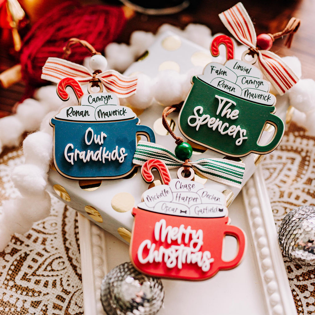 Three Christmas hot cocoa personalized family ornaments are displayed on a white tray with holiday decorations. T