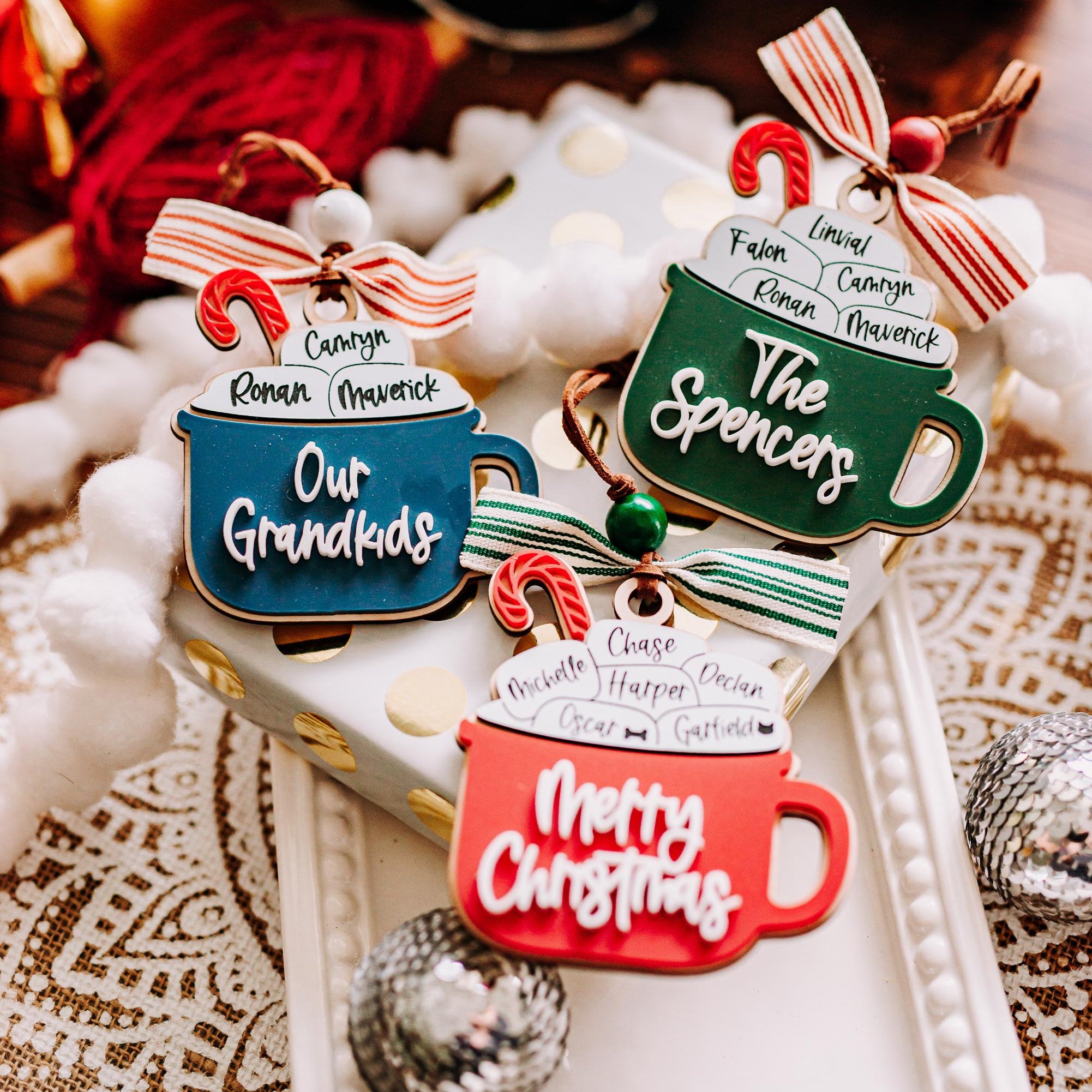 Three Christmas hot cocoa personalized family ornaments are displayed on a white tray with holiday decorations. T