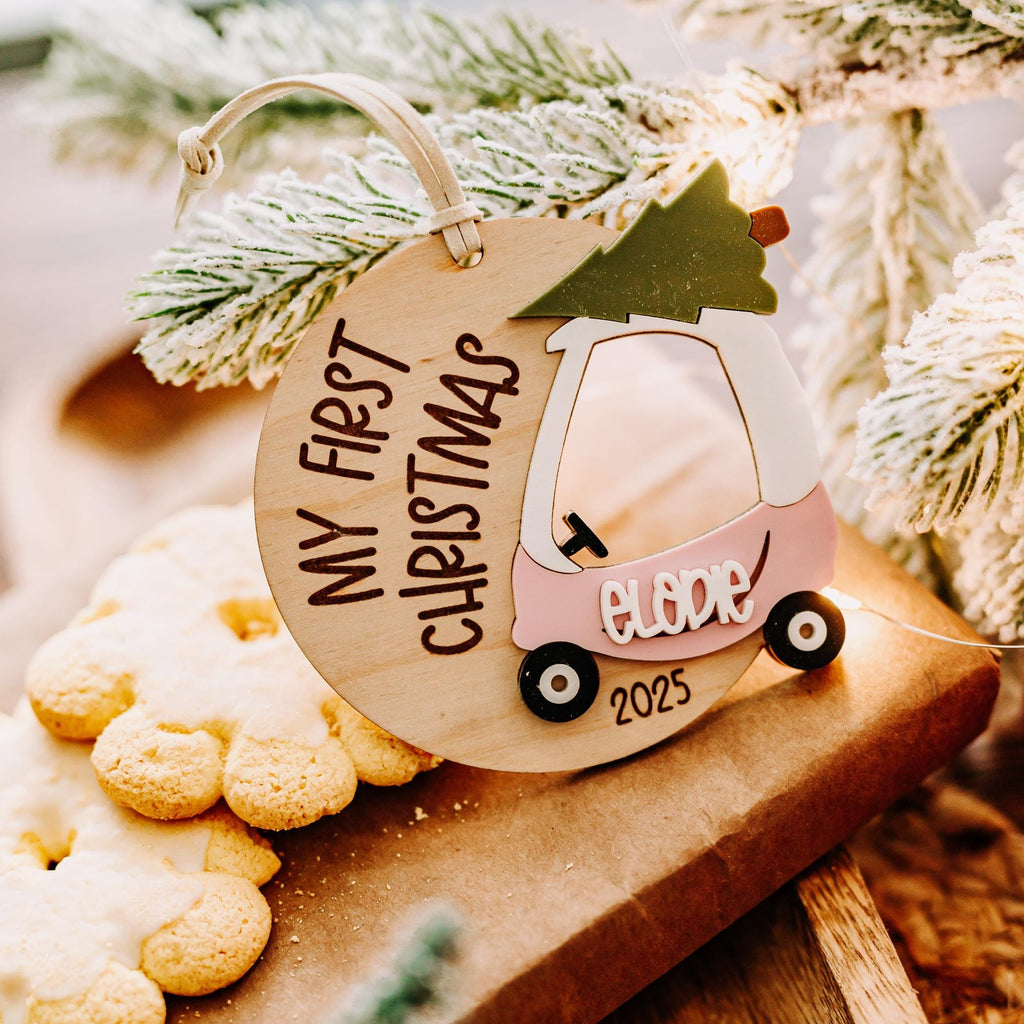 A personalized wooden Christmas ornament shaped like a pink and cozy coupe car with a green tree on top, hanging on a wooden board with cookies and frosted pine branches.