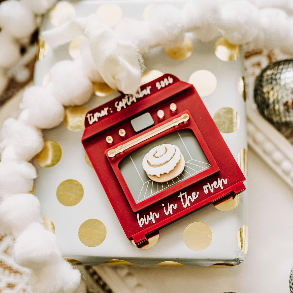 A Christmas ornament shaped like a retro oven with a small "bun in the oven", placed on top of a white gift box with gold polka dots and surrounded by white pom-pom garland and silver ornaments.