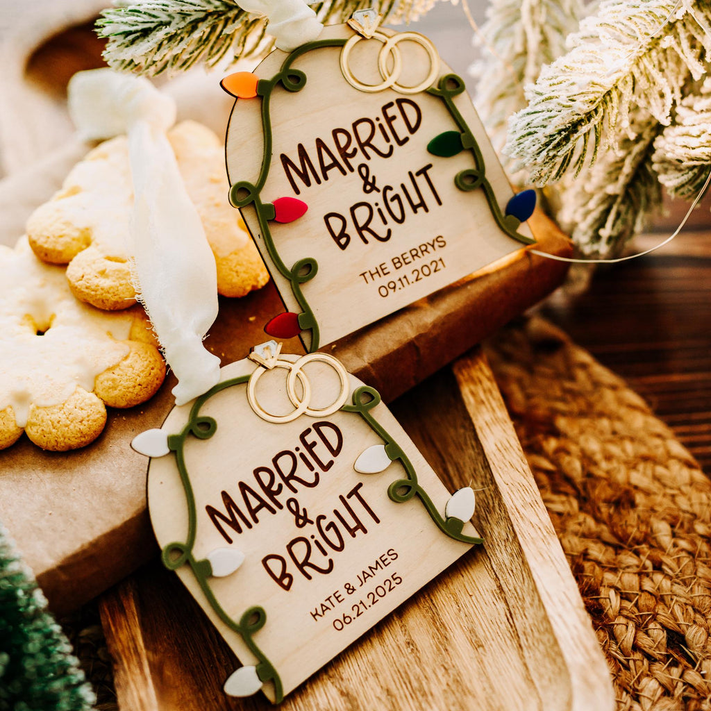 Two personalized Christmas ornament tags reading 'MARRIED & BRIGHT' with gold wedding rings printed at the top, styled on a wooden board with cookies, evergreen branches, and a woven textile.
