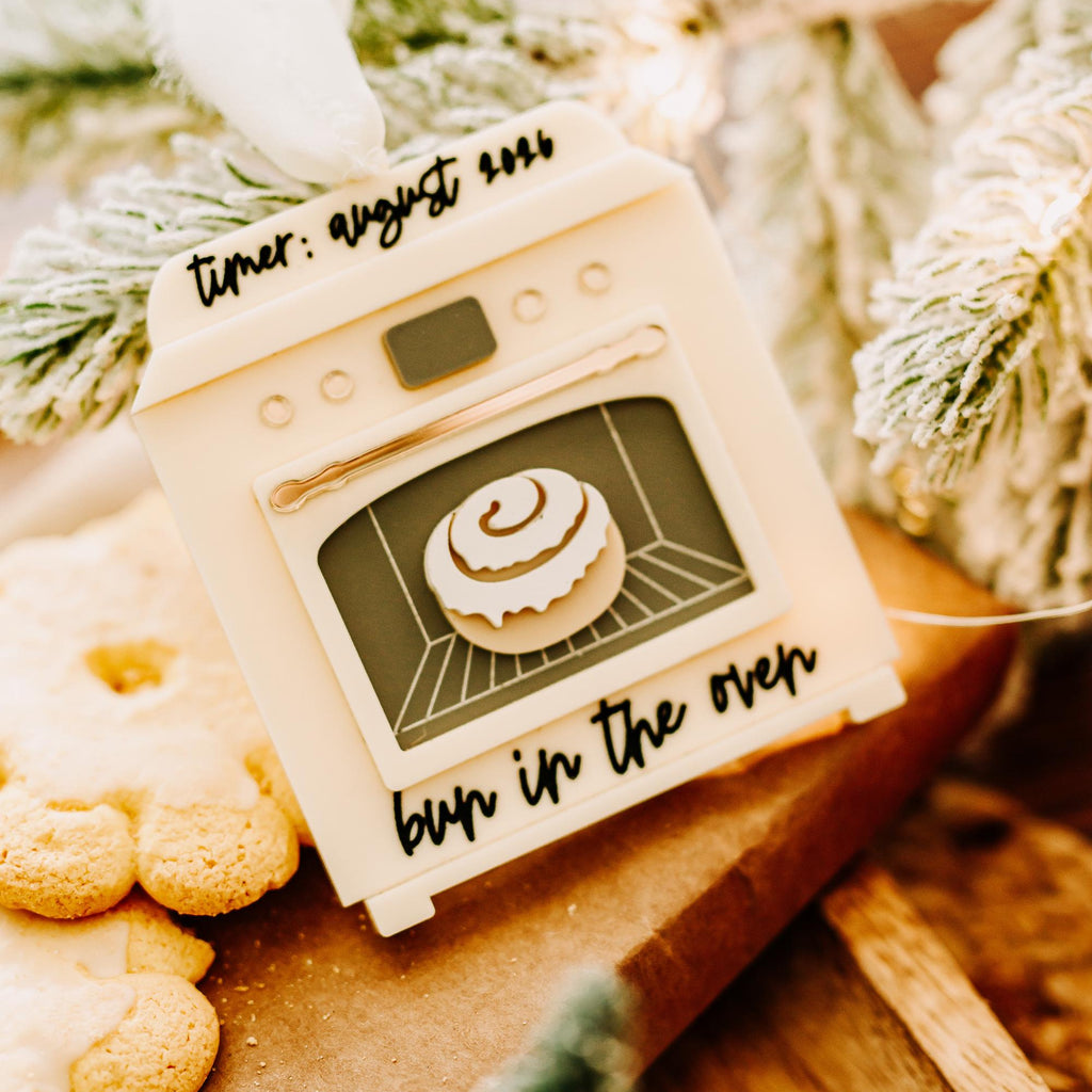 A decorative oven-shaped ornament  a cinnamon roll illustration and the words 'timer: august 2019' and 'burn in the over,' styled on a wooden surface with cookies and frosted greenery in the background.