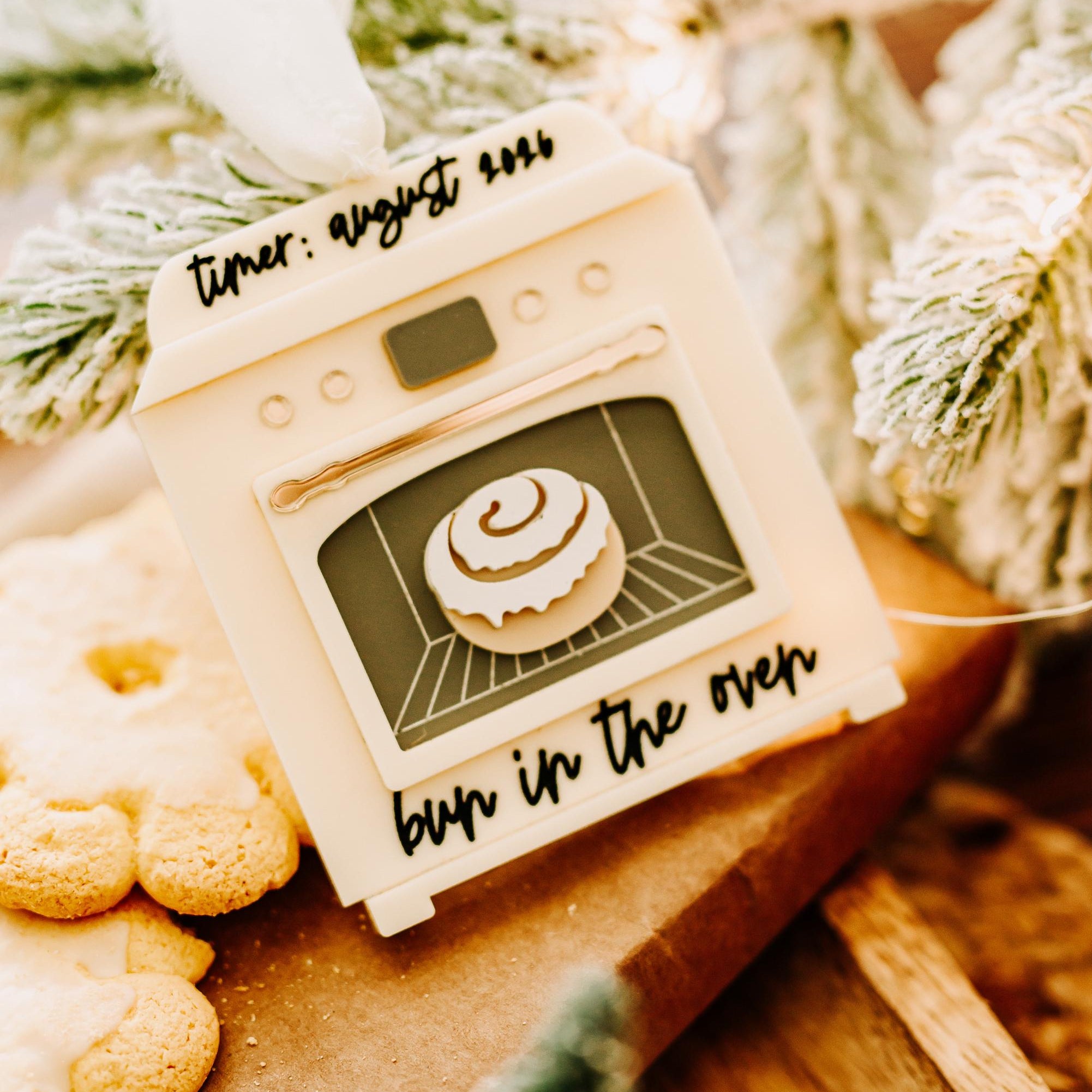 A decorative oven-shaped ornament  a cinnamon roll illustration and the words 'timer: august 2019' and 'burn in the over,' styled on a wooden surface with cookies and frosted greenery in the background.