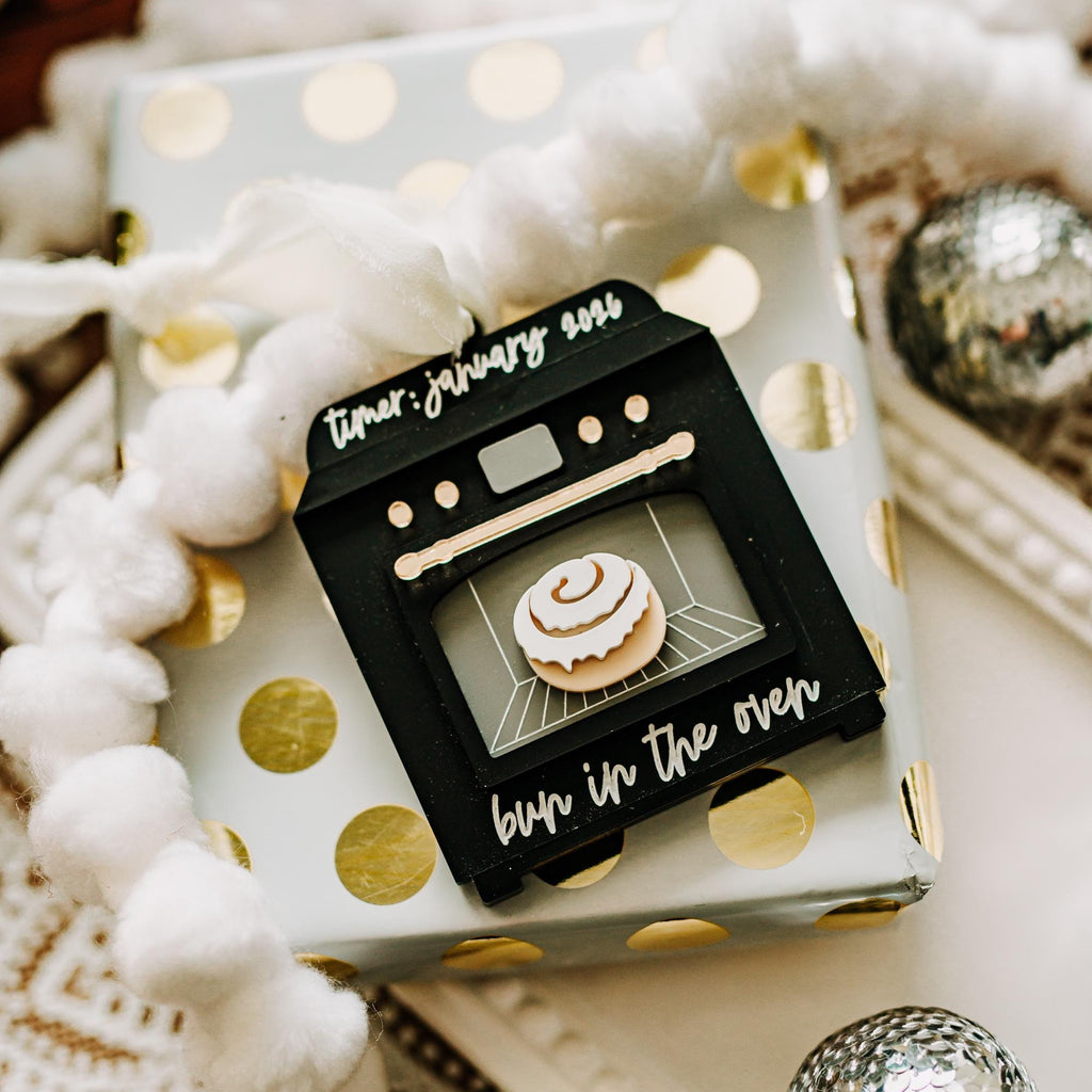 A small black oven-shaped ornament or decorative piece with a clear window showing a tiny cinnamon roll, placed on top of a white gift box with gold polka dots. The scene includes white pom-pom garland, a patterned textile, and silver decorative balls in a styled setting.