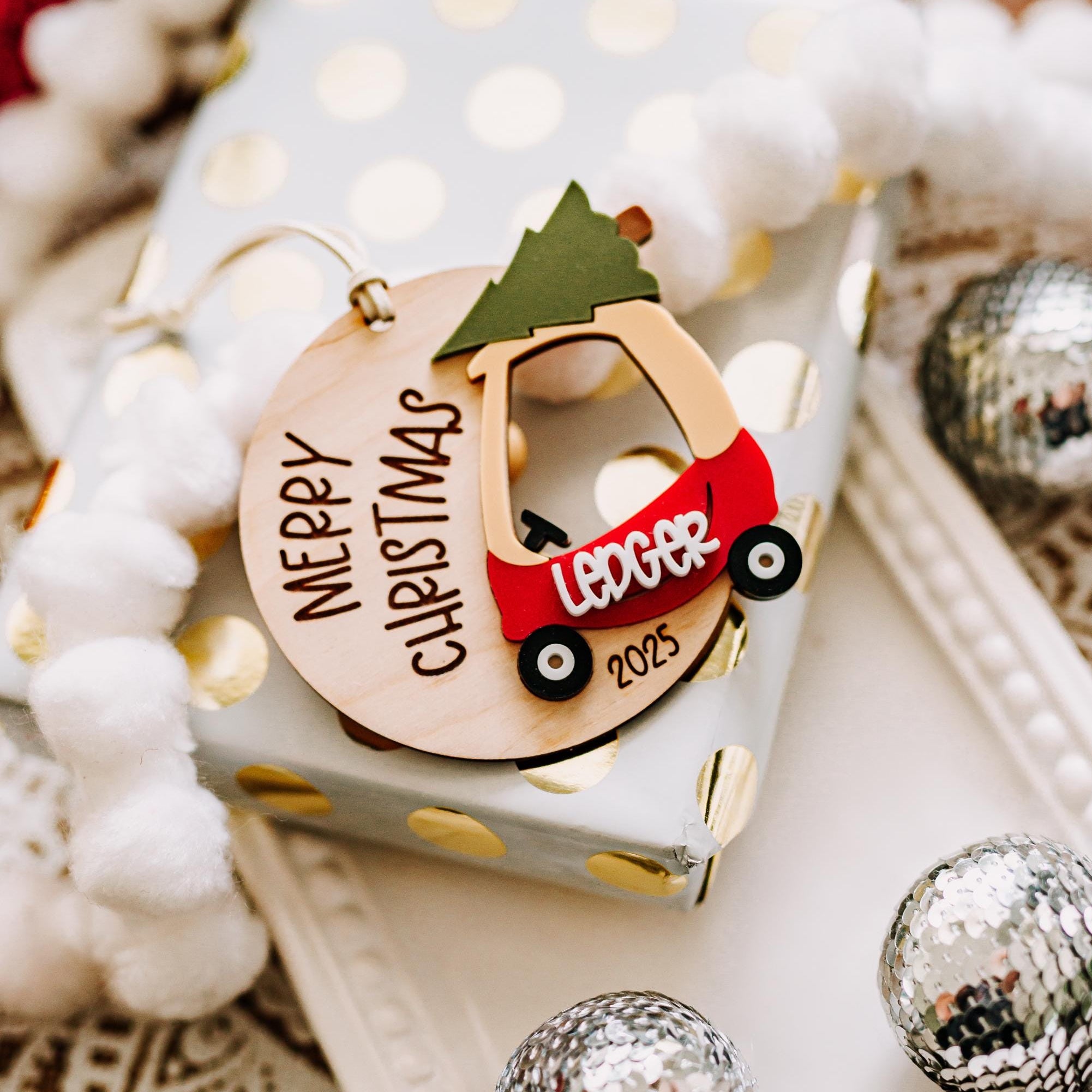 A personalized Christmas ornament shaped like a red cozy coupe carrying a green Christmas tree, with the text 'Merry Christmas 2025' written on a light wood circle. The ornament is styled on a white gift box with gold polka dots, surrounded by white fluffy garland and silver holiday ornaments.