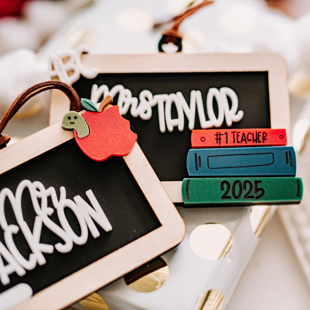Personalized teacher-themed ornament decorations featuring chalkboard-style tags with names, a red apple accent, and a stack of colorful books labeled #1 TEACHER and 2025, styled on a white surface with bokeh lights in the background.
