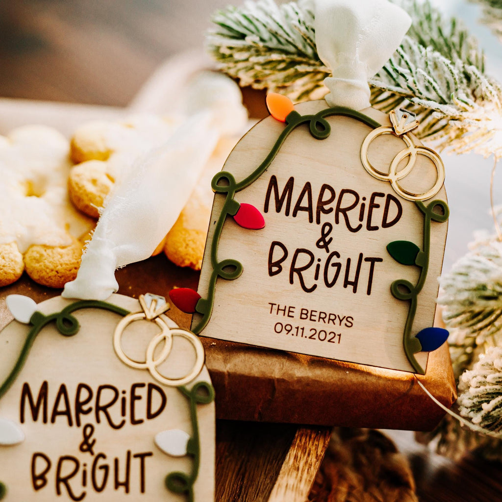 Personalized wooden Christmas ornament tags with the text 'MARRIED & BRIGHT' and wedding ring illustrations, styled with flocked pine branches and cookies on a rustic wooden surface.