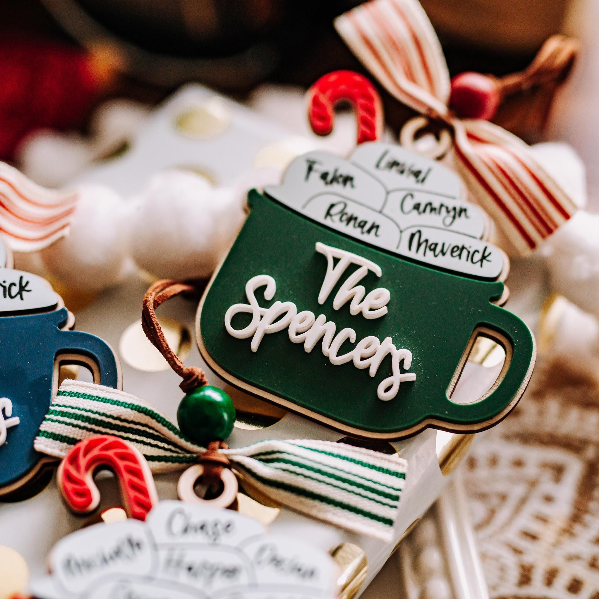 A close-up of several Christmas ornaments shaped like hot cocoa mugs, each with white script names written on them, arranged with candy canes and ribbons on a soft holiday background.