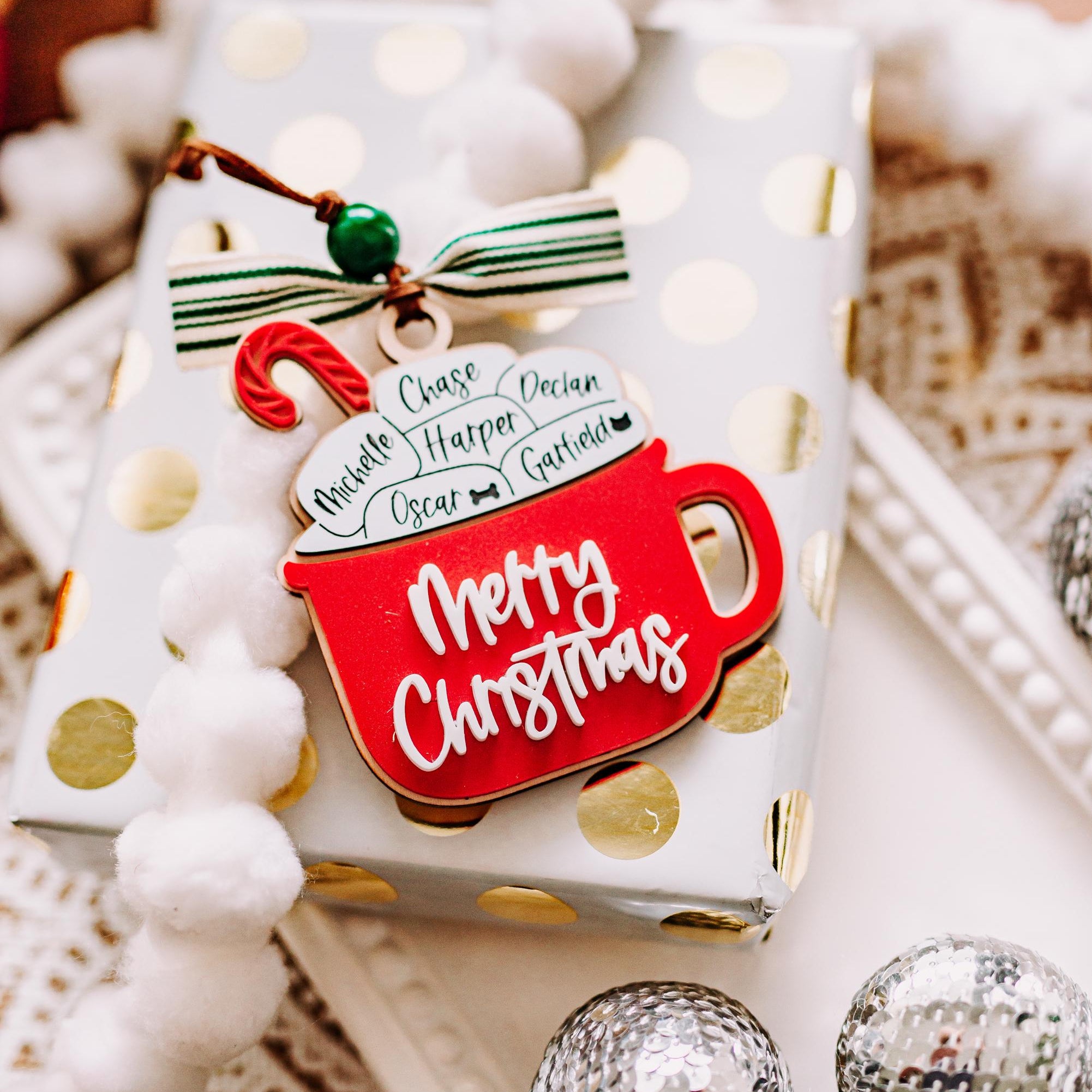 A festive Christmasornament shaped like a red mug with the words 'Merry Christmas' in white script, attached to a wrapped gift with gold polka dots. The tag includes a small hanging ornament with a candy cane and a green-and-white striped ribbon.