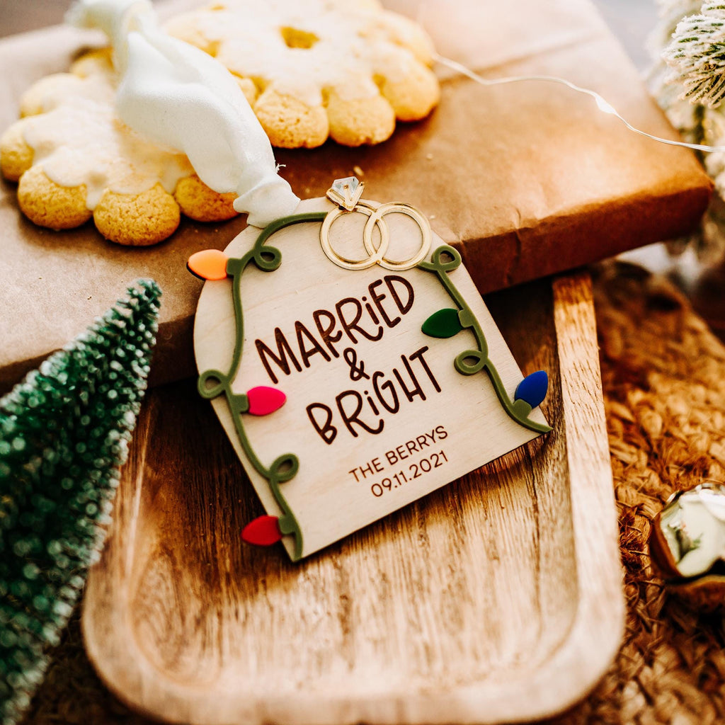 A personalized Christmas ornament tag hanging from a wrapped gift on a wooden tray. The tag reads 'MARRIED & BRIGHT' with the date '09.11.2021'