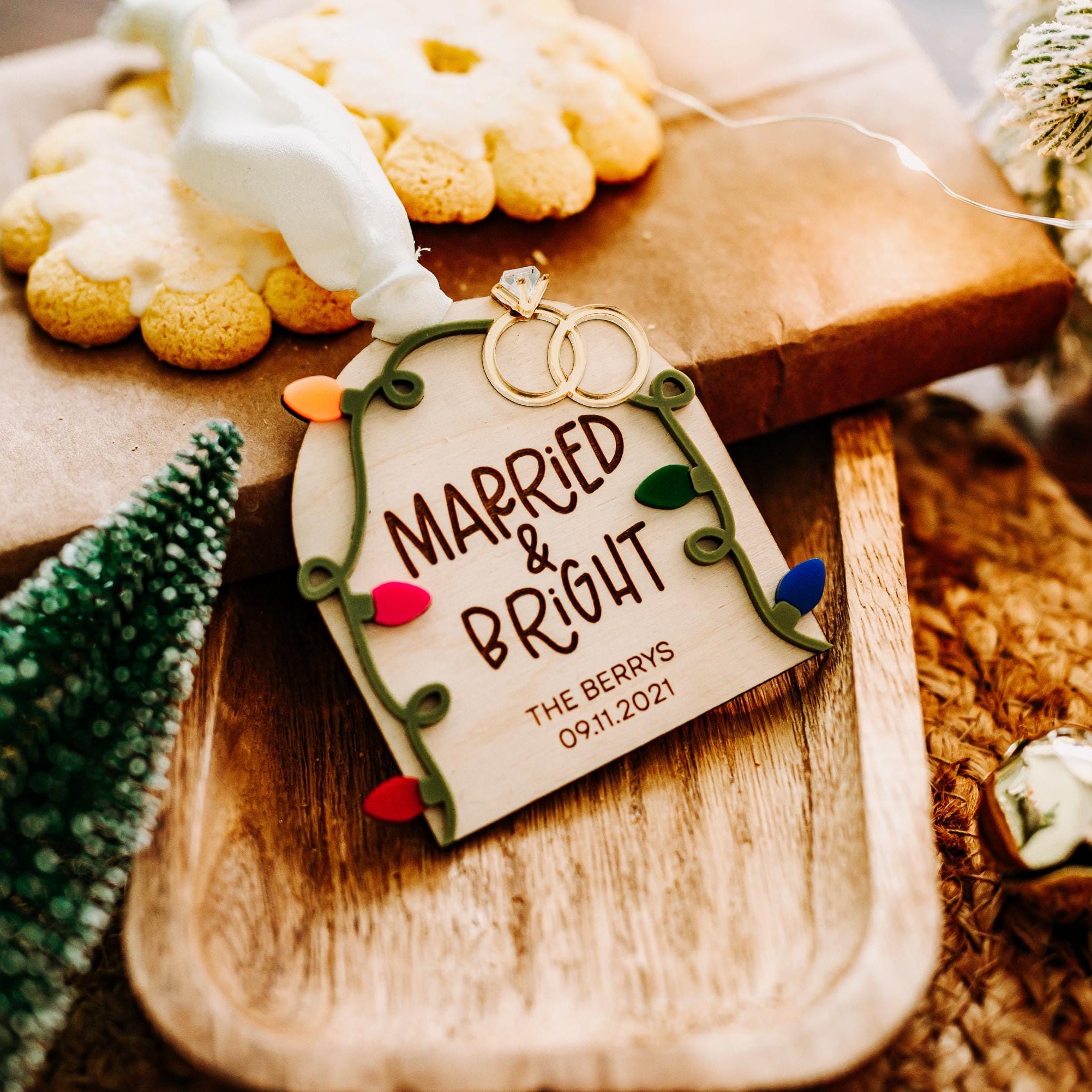 A personalized Christmas ornament tag hanging from a wrapped gift on a wooden tray. The tag reads 'MARRIED & BRIGHT' with the date '09.11.2021'