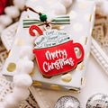 A wrapped gift box with white and gold polka dot paper, decorated with a red mug-shaped Christmas ornament that says 'Merry Christmas' and a hot cocoa  mug ornament reading 'Merry Christmas , with the names Harper, Declan, Oscar, Garfield, Michelle, & Chase' on the hot cocoa marshmallows. The box is styled with white pom-pom garlands, silver ornaments, and a red ball on a beige textured surface.