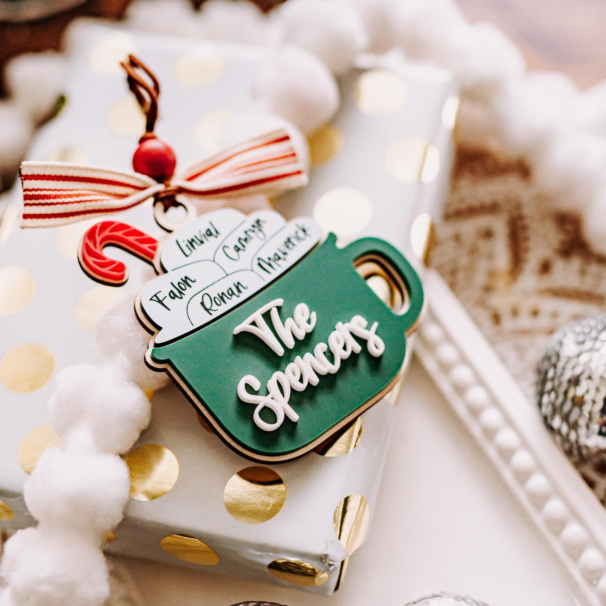 A close-up of a wrapped gift box with a green hot cocoas mug shaped ornament with 'The Spencer' and a red-and-white striped bow, styled with white pom-pom beads and silver mirrored ornaments on a beige surface.