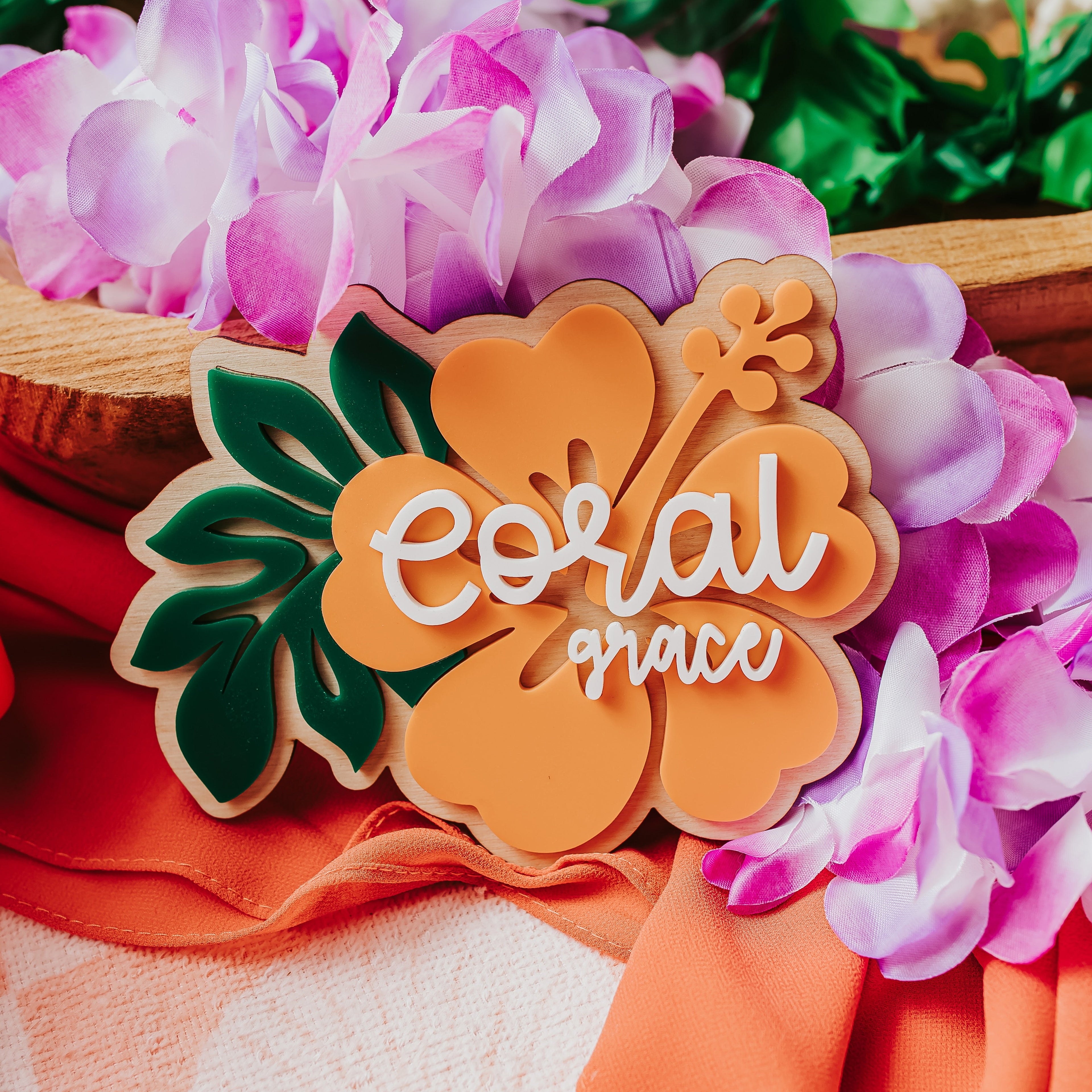 A decorative wooden baby name sign shaped like a hibiscus flower with the words 'coral grace' in white script, styled with pink flowers, green leaves, orange fabric, and a peach towel.