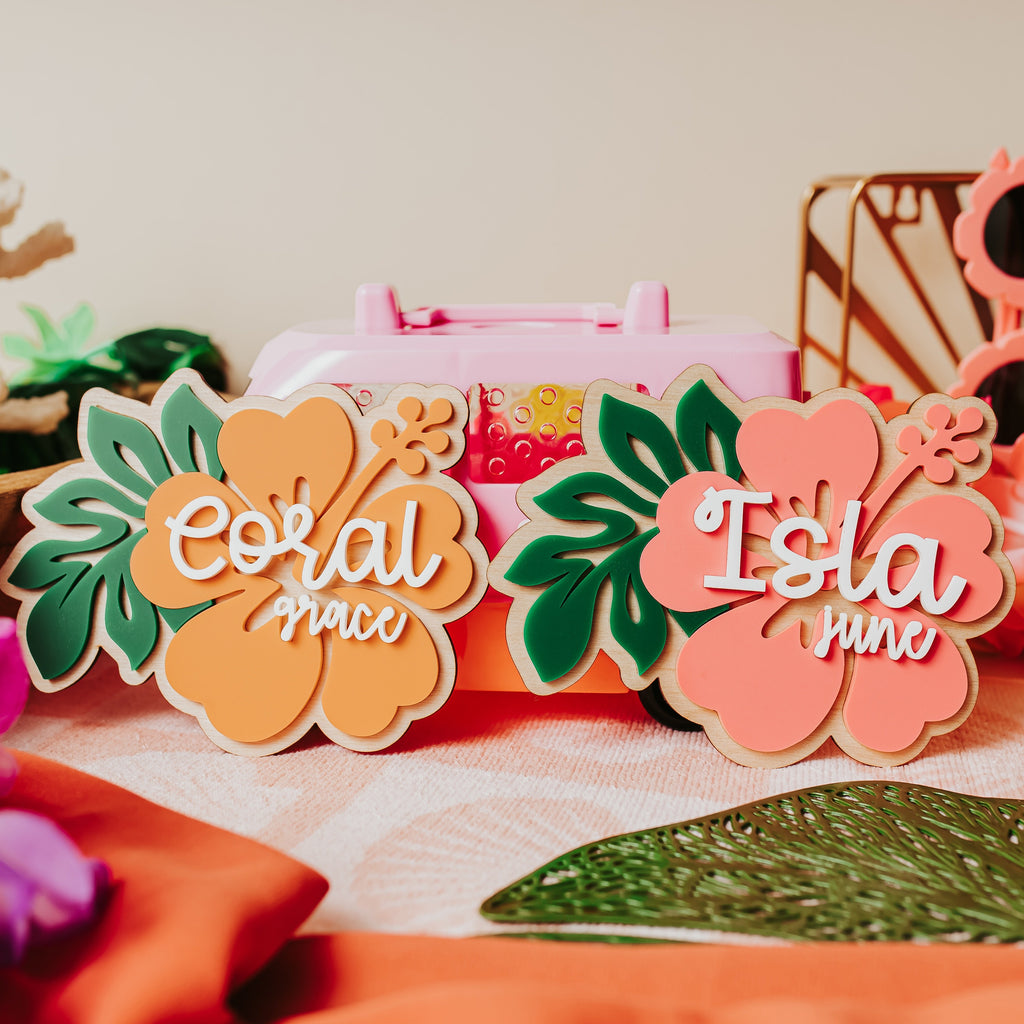 Two tropical hibiscus flower-shaped acrylic baby name signs reading 'Coral grace' and 'Isla junre' displayed on a table with tropical props including flowers, sunglasses, and a pink case.