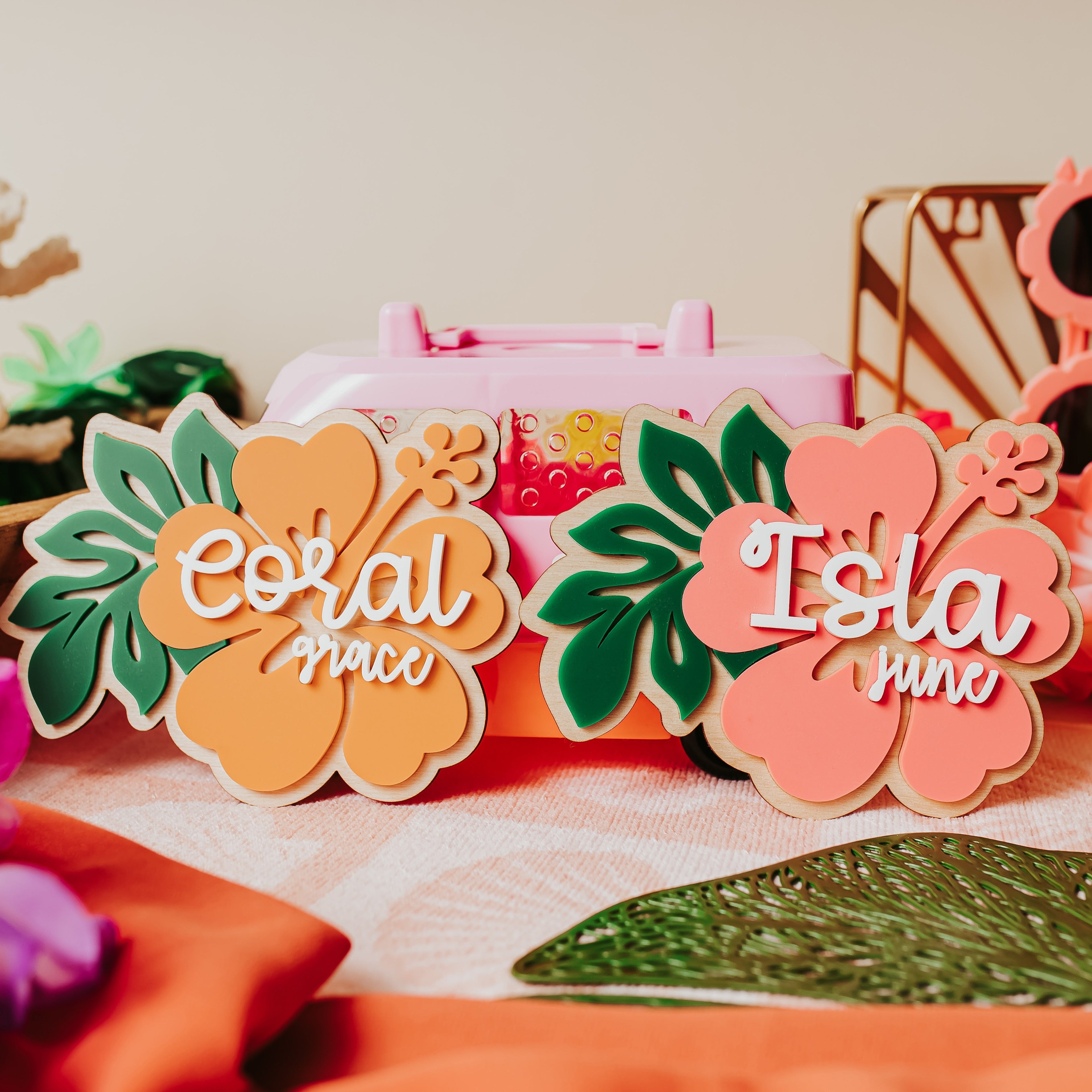 Two tropical hibiscus flower-shaped acrylic baby name signs reading 'Coral grace' and 'Isla junre' displayed on a table with tropical props including flowers, sunglasses, and a pink case.