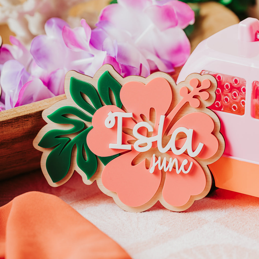 A coral-pink wood and acrylic baby name sign shaped like a hibiscus flower with green leaves and the words 'Isla June' in white script, styled on a table with tropical flowers and a pink suitcase in the background.