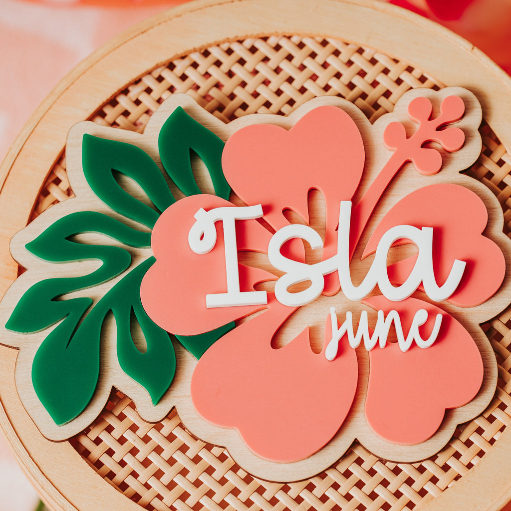 A close-up of a wood and acrylic hibiscus tropical flower and leaf baby namr sign in coral pink, green, and cream, with the words 'Isla june' in white script across the center.