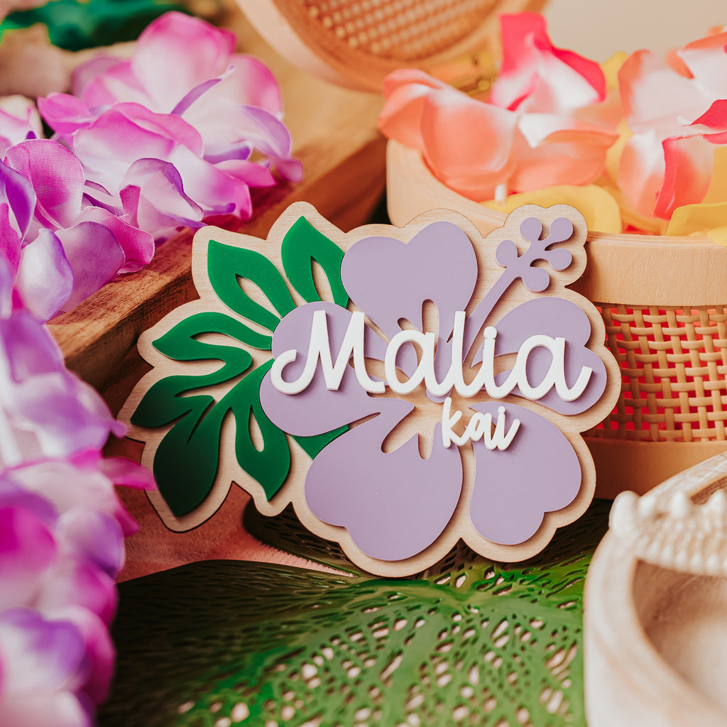A decorative wooden and acrylic baby name announcement sign with a hibiscus flower design and the names 'Malia kai' in white script, styled with tropical flowers and woven baskets.