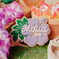 A decorative wooden and acrylic baby name announcement sign with a hibiscus flower design and the names 'Malia kai' in white script, styled with tropical flowers and woven baskets.