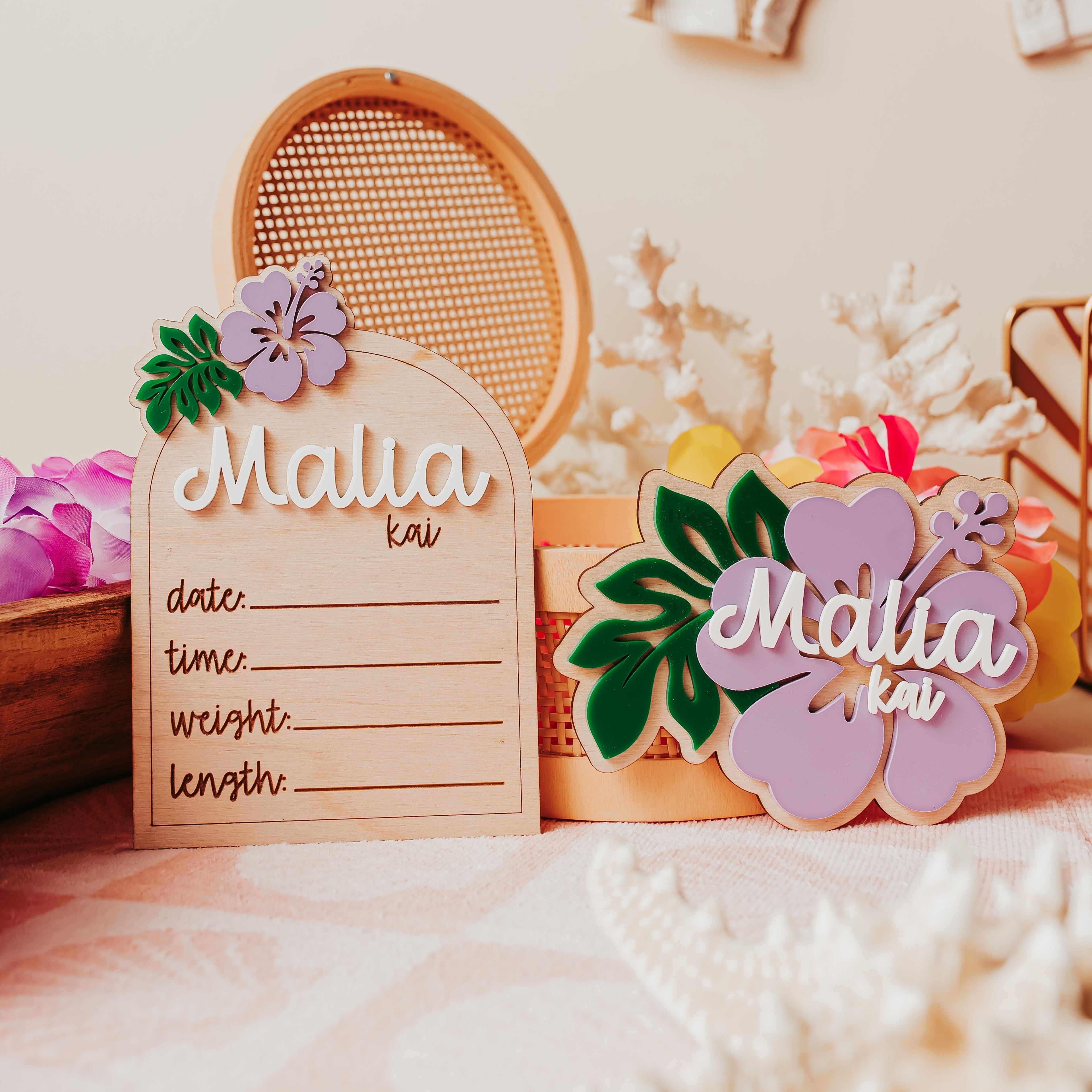 A styled tabletop display featuring a wooden arch-shaped sign engraved with 'Malia kai' and lined for writing, alongside matching wooden hibiscus-shaped  with floral accents, a woven oval tray, and soft beach-themed decor in a warm neutral setting.