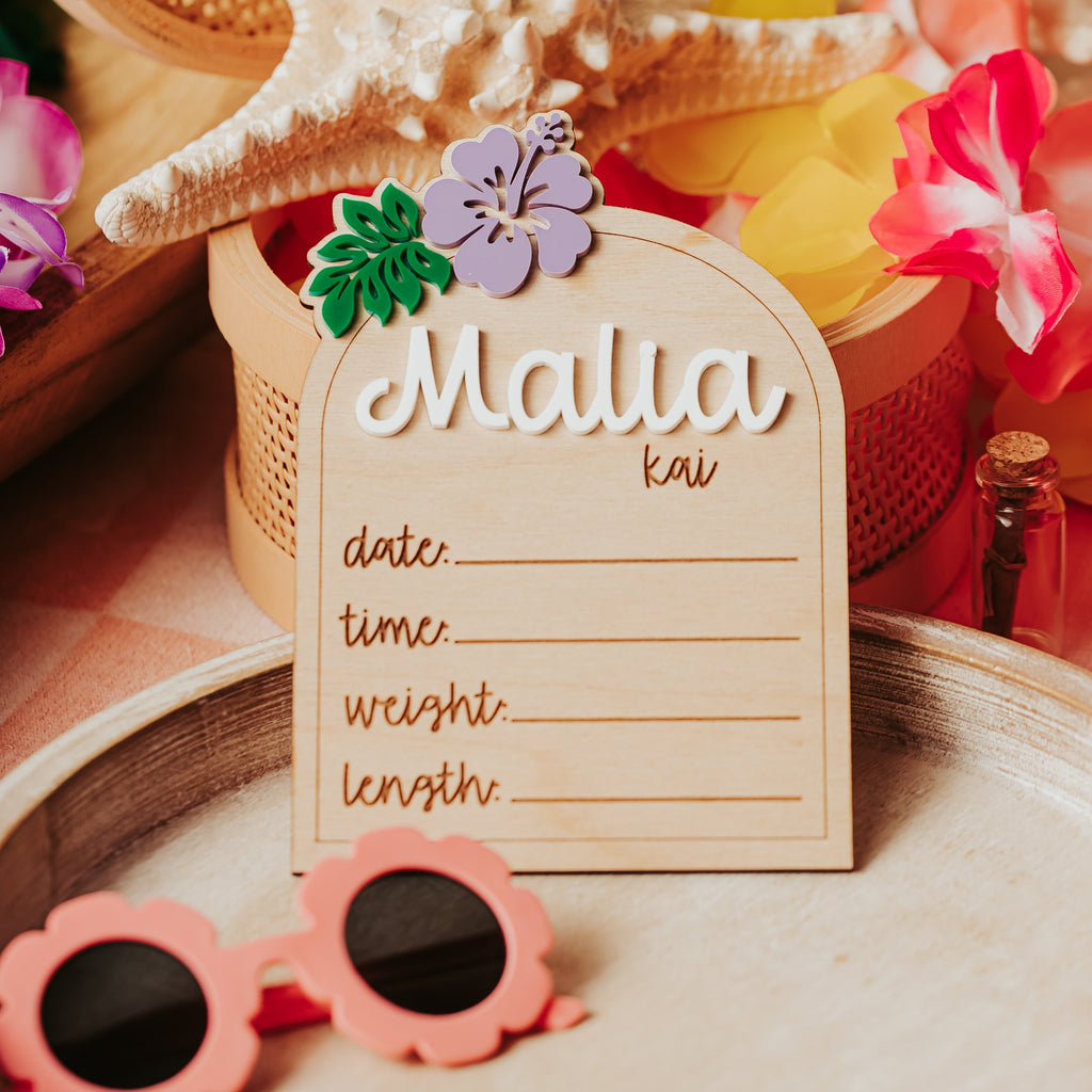 A wooden arch-shaped beach-themed baby stat sign reading 'Malia kai' with lines for date, time, weight, and length, styled with a starfish, woven basket, tropical flowers, and pink sunglasses on a tray.