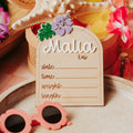 A wooden arch-shaped beach-themed baby stat sign reading 'Malia kai' with lines for date, time, weight, and length, styled with a starfish, woven basket, tropical flowers, and pink sunglasses on a tray.