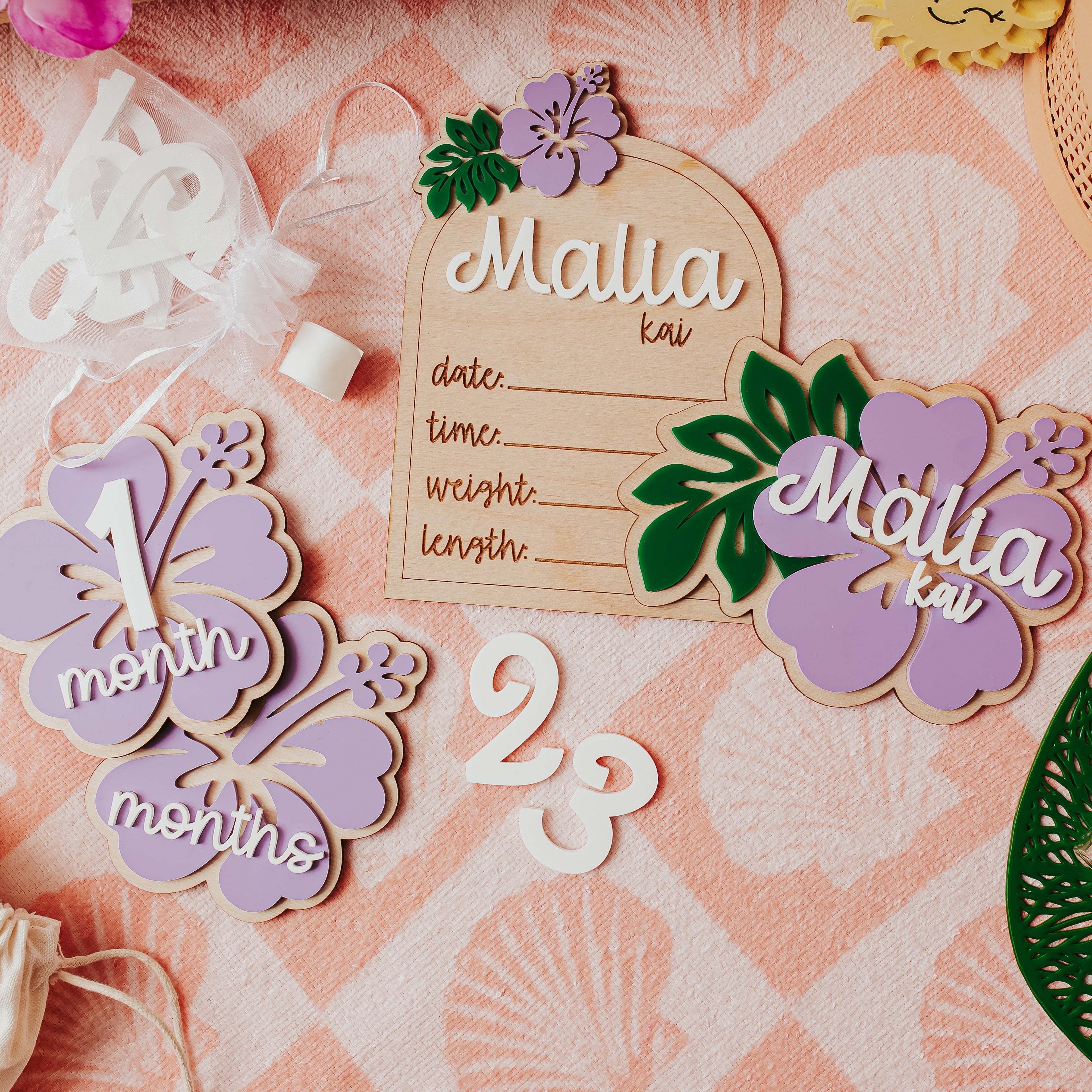 A styled flat-lay of personalized wooden baby milestone plaquesm baby name announcement and birth stat on a pink patterned fabric background. The set includes a wooden birth date plaque, a lavender '1 month' hibiscus flower milestone, a months' flower milestone, and a lavender 'Malia kai' name plaque with green leaves.