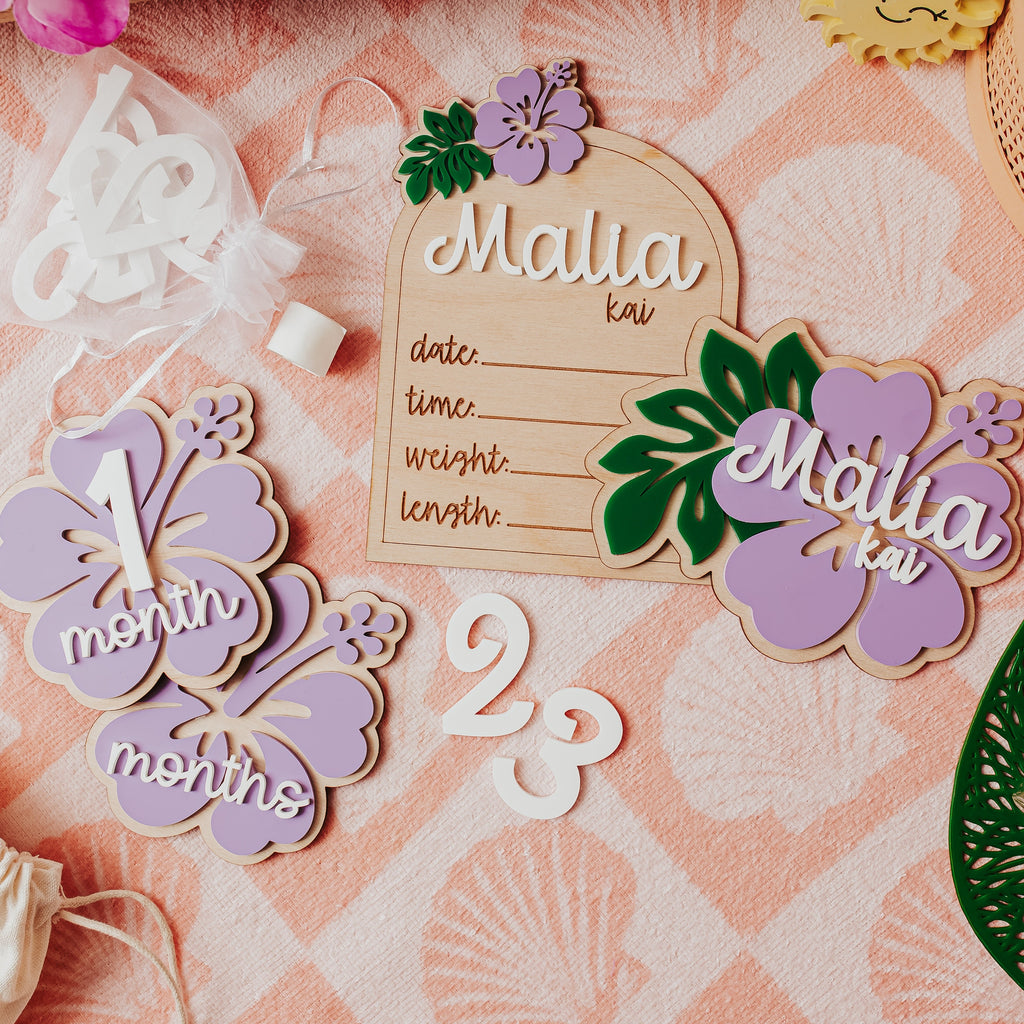 A styled flat-lay of personalized wooden and acrylic baby milestone plaques , birth stat and name sign on a pink patterned fabric background. The set includes a wooden birth date plaque, a lavender '1 month' flower milestone, a lavender '23 months' flower milestone, and a lavender 'Malia kai' name plaque with green leaves.