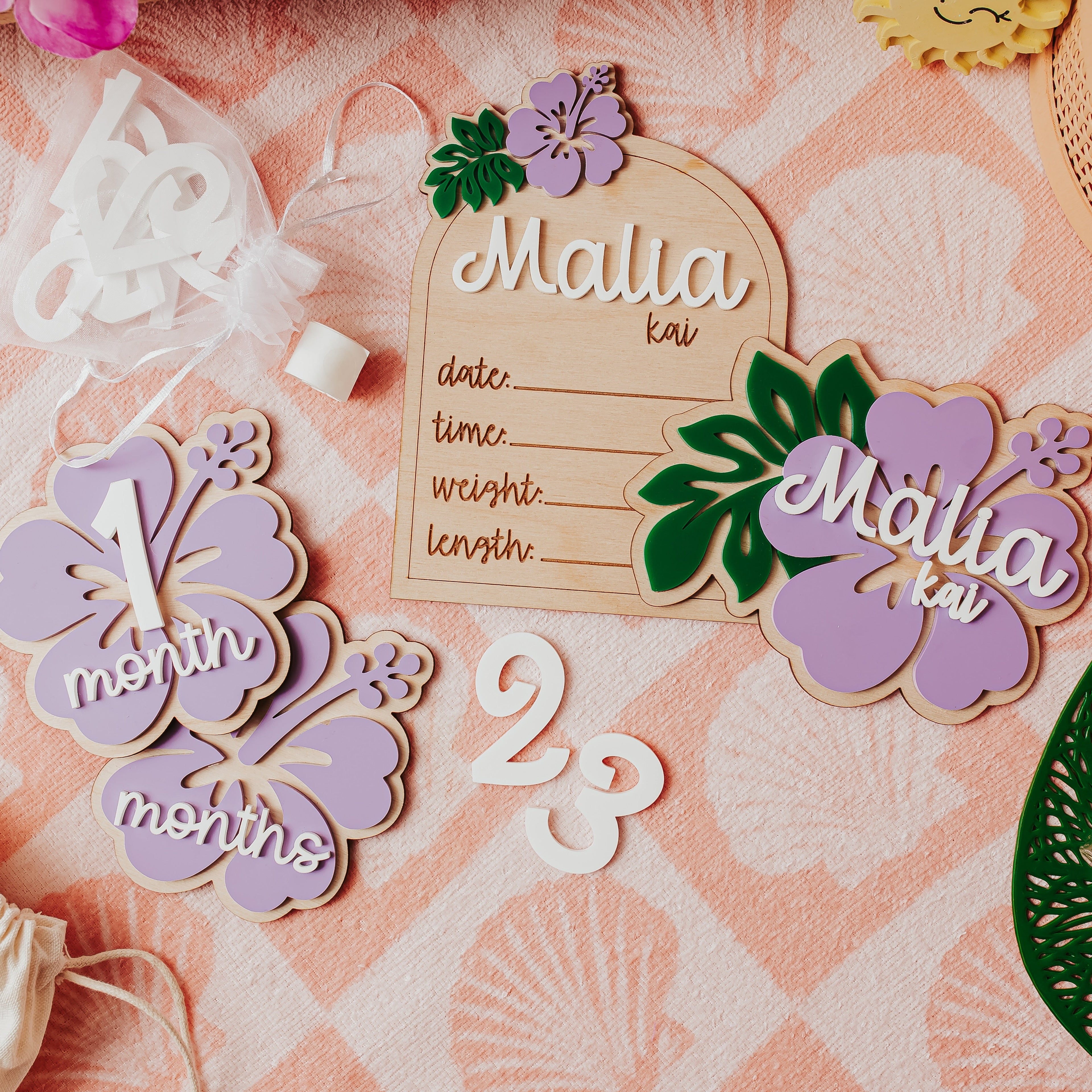 A styled flat-lay of personalized wooden and acrylic baby milestone plaques , birth stat and name sign on a pink patterned fabric background. The set includes a wooden birth date plaque, a lavender '1 month' flower milestone, a lavender '23 months' flower milestone, and a lavender 'Malia kai' name plaque with green leaves.