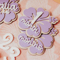 Close-up of decorative acrylic and wooden milestone plaques shaped like hibiscus flowers, one reading 'Malia kai' with green leaves and the other reading '1 month' and 'months' in white script, arranged on a peach patterned surface.