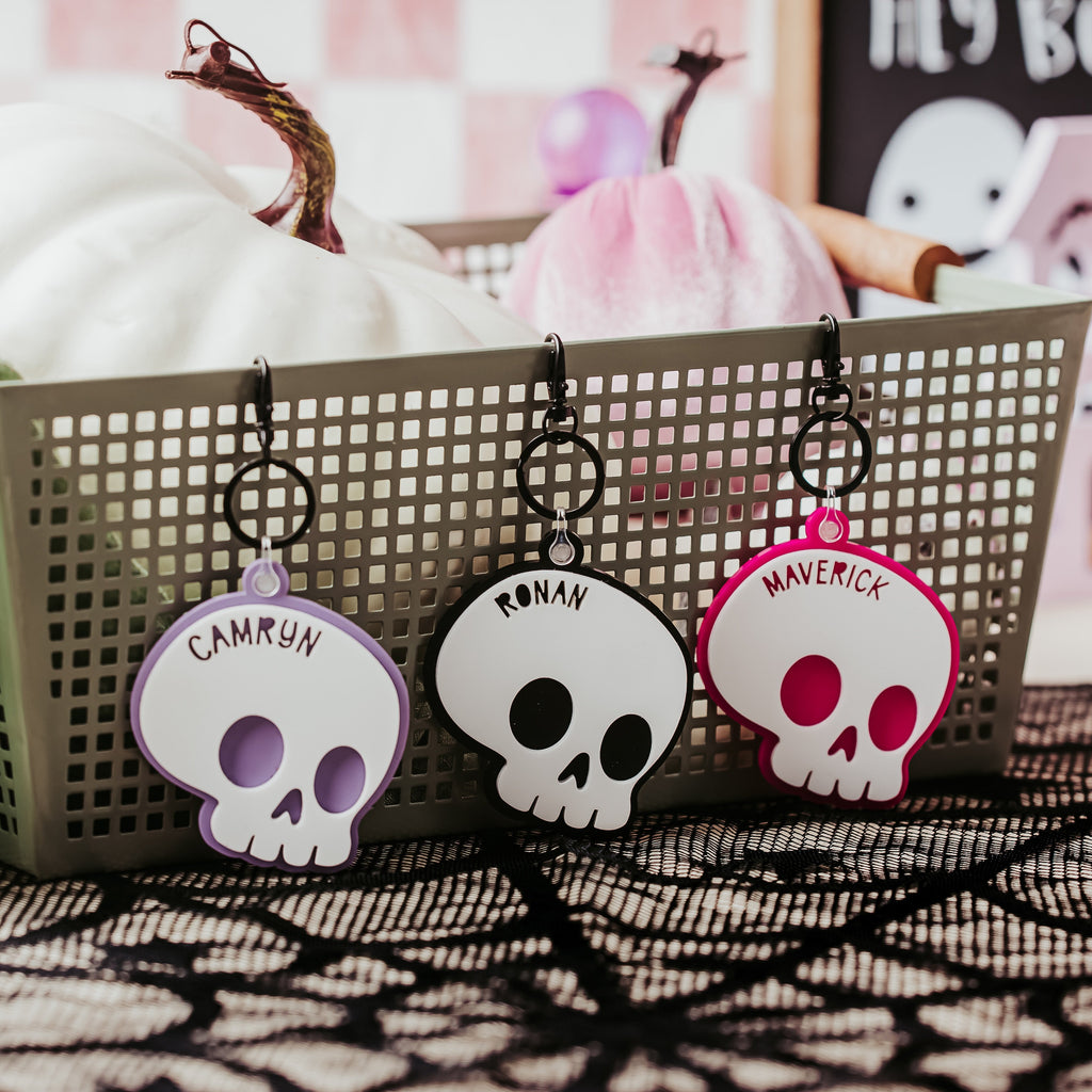 Three Halloween-themed skull keychains hanging from a gray plastic basket on a black lace surface, with pumpkins and a ghost decoration in the background.