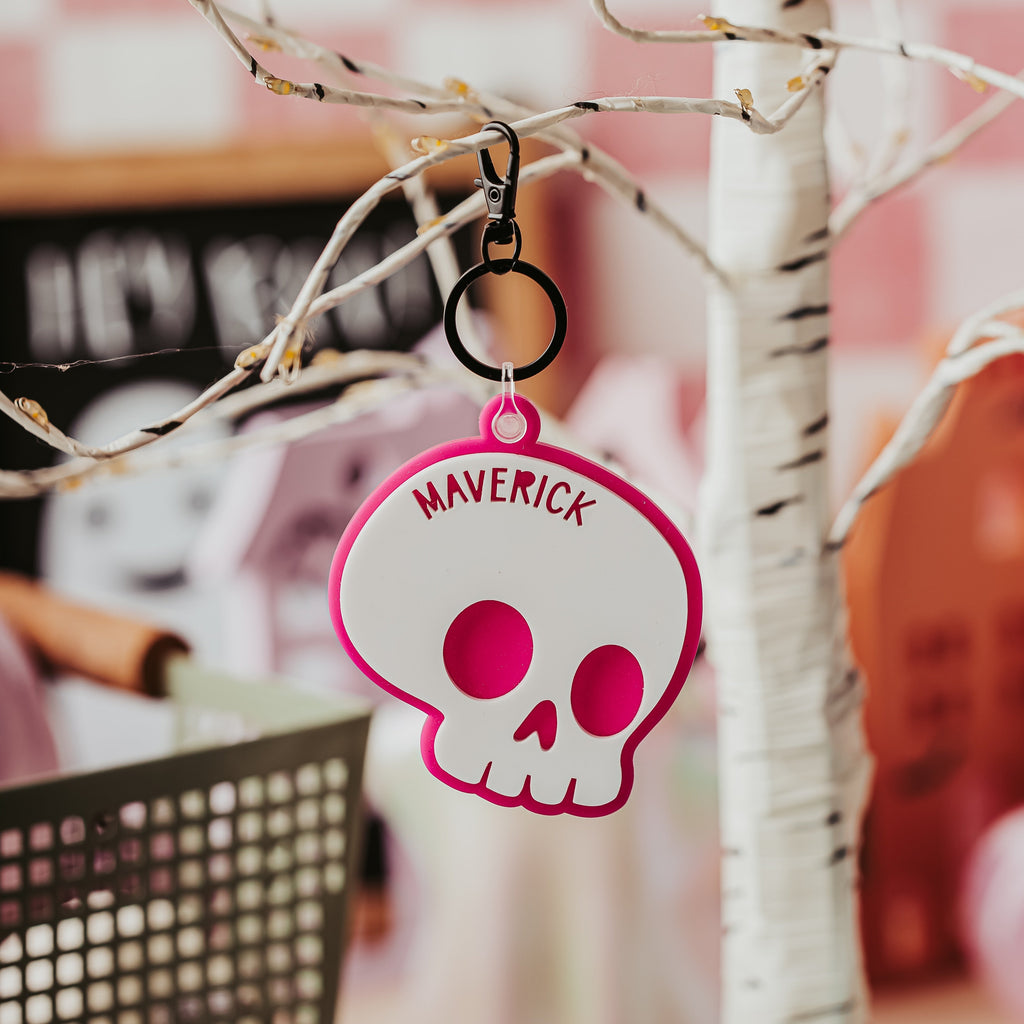 A white skull-shaped keychain or bag charm with a hot pink border and the name 'MAVERICK' printed in pink, hanging from a black clip on a decorative tree branch.