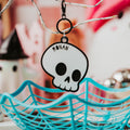 A Halloween-themed skull keychain with the name 'RONAN' printed on it, hanging from a decorative branch above a turquoise spiderweb basket.