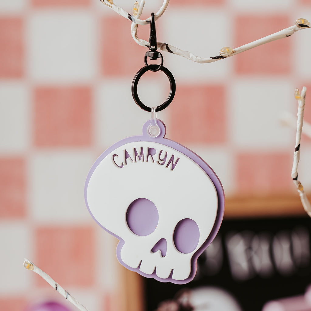 A custom Halloween basket tag shaped like a skull, featuring a white face with a lavender-purple border and the name 'CAMRYN' printed on the forehead. The plaque hangs from a black ring clip attached to a decorative branch, set against a soft pink and white checkered background.