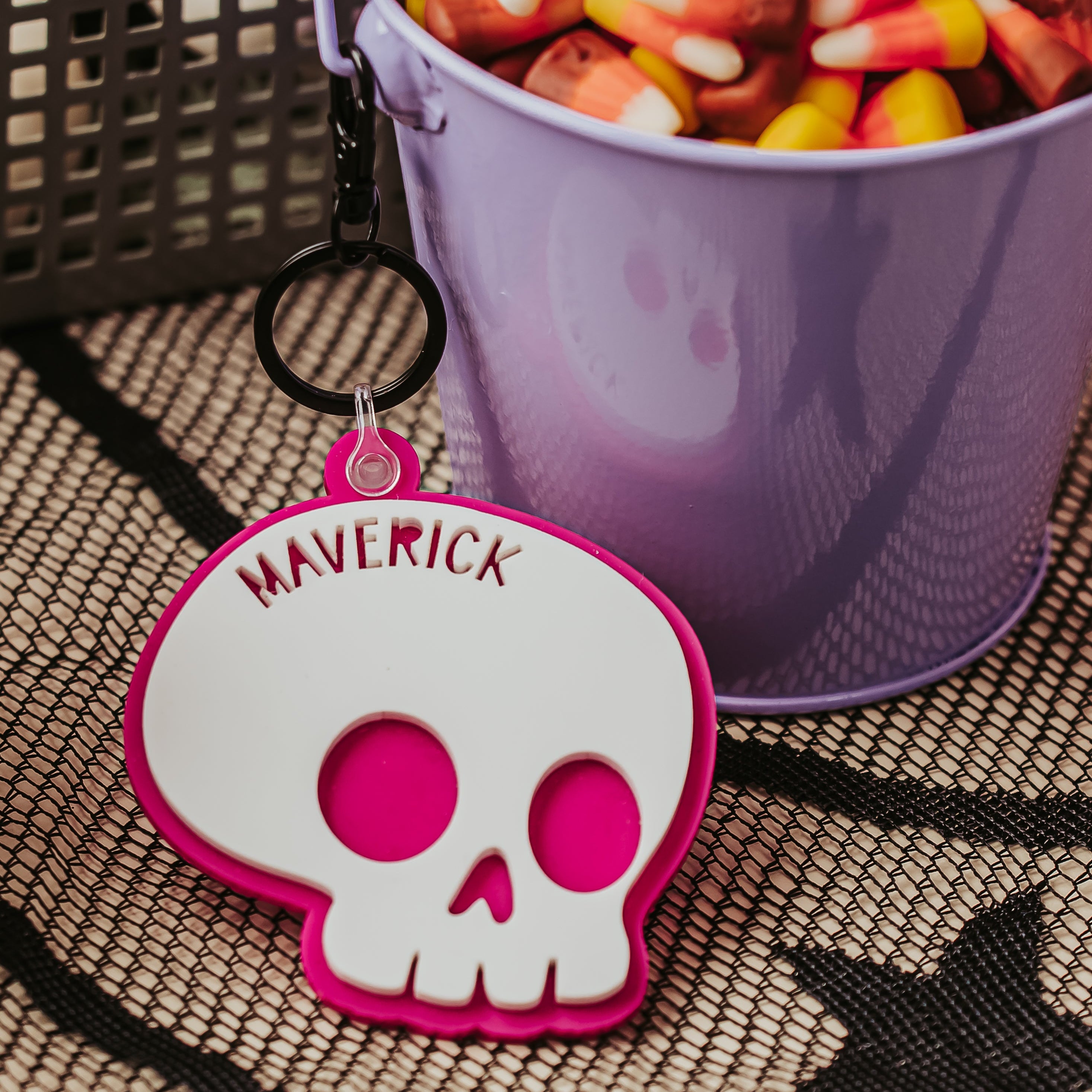 A Halloween-themed product photo showing a custom skull keychain with the name 'MAVERICK' in hot pink, next to a purple bucket filled with candy corn.