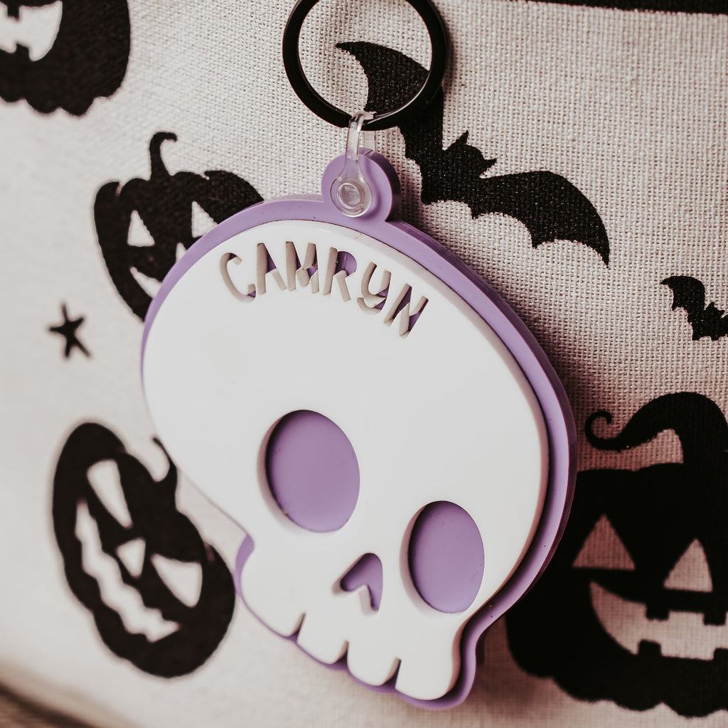 A Halloween-themed fabric storage basket with a black rim, decorated with black pumpkins, bats, and stars on an off-white background. A white and lavender skull-shaped tag with the name 'CAMRON' is attached to a black plastic ring.