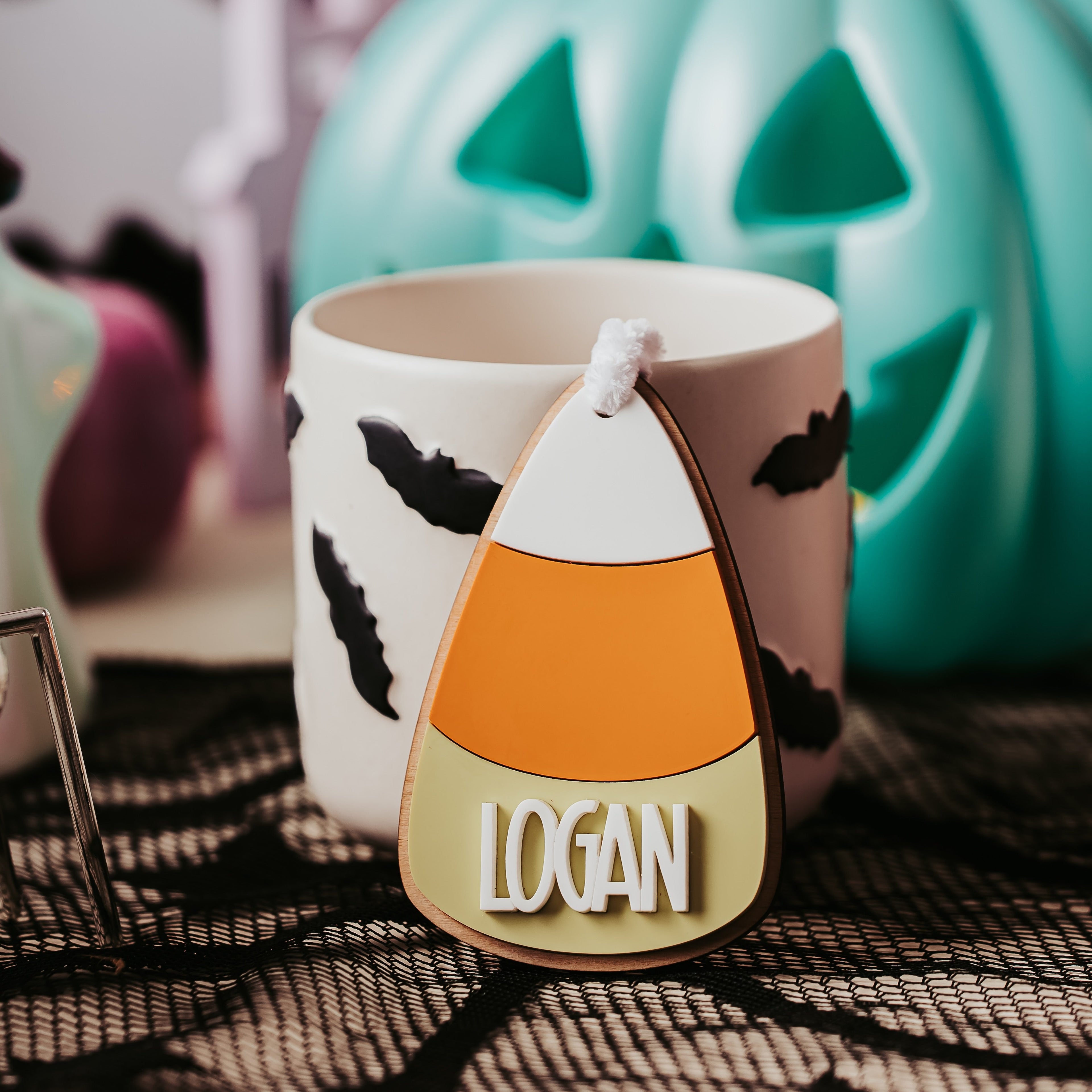 A white ceramic mug with black bat silhouettes, featuring a candy corn-shaped boo basket tag that reads 'LOGAN' in white letters. The mug is styled on a black spiderweb fabric with a large teal jack-o'-lantern and other Halloween decorations blurred in the background.