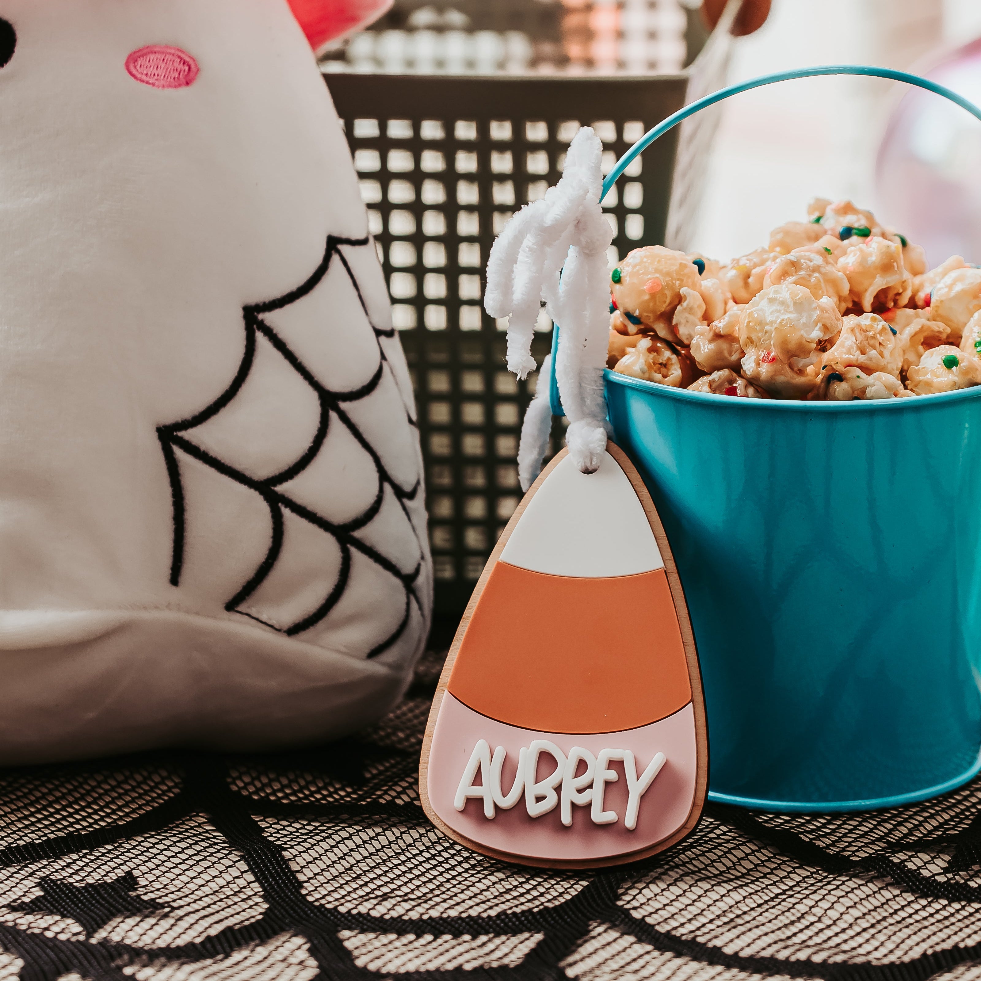 A Halloween-themed product display featuring a white plush ghost toy with a pink hat, a turquoise bucket filled with popcorn, and a candy corn-shaped boo basket tag personalized with the name 'AUBREY' on a patterned rug.