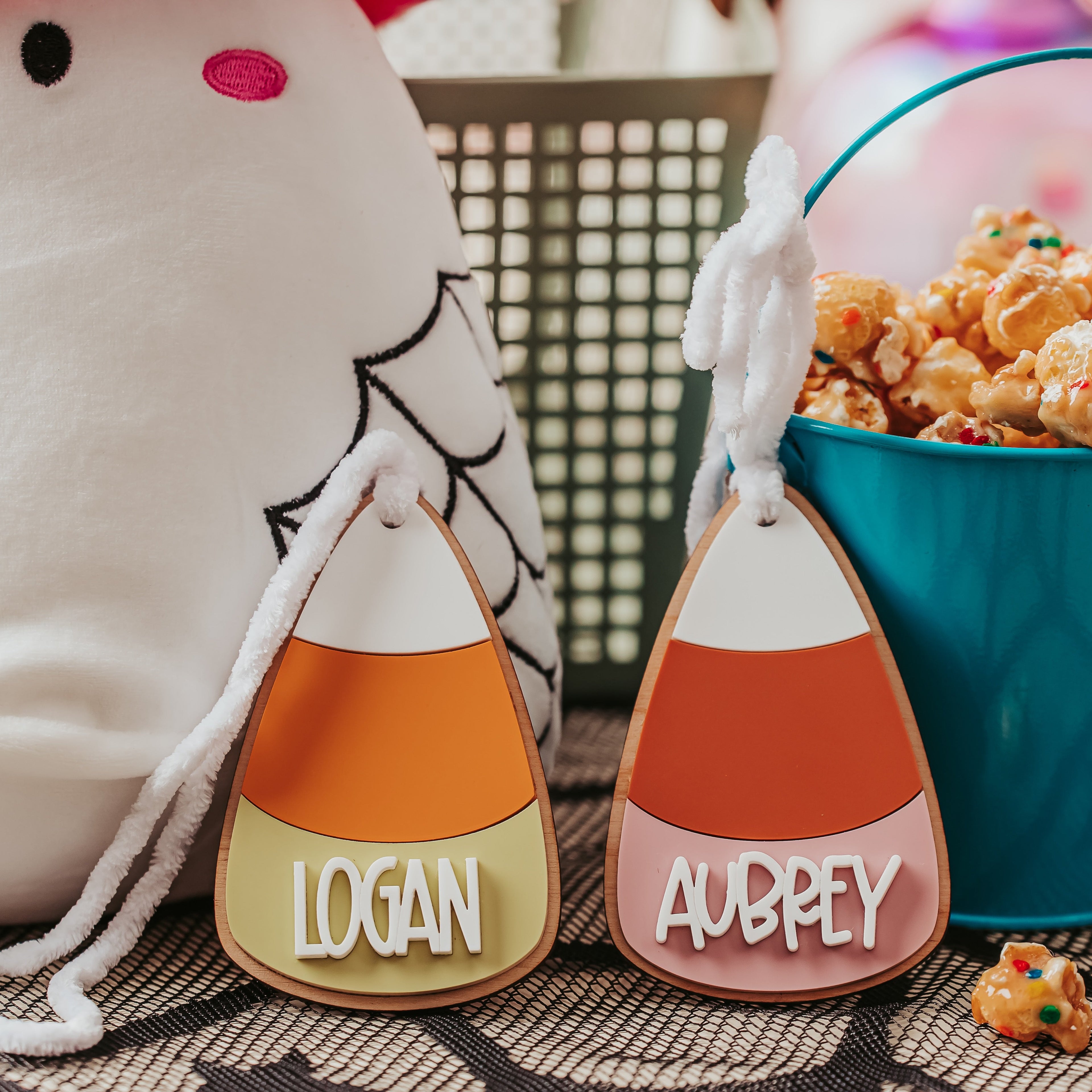 Personalized candy corn boo basket tag with the names 'LOGAN' and 'AUBREY' in white lettering, shown next to a teal bucket of popcorn and a white plush toy at a party.