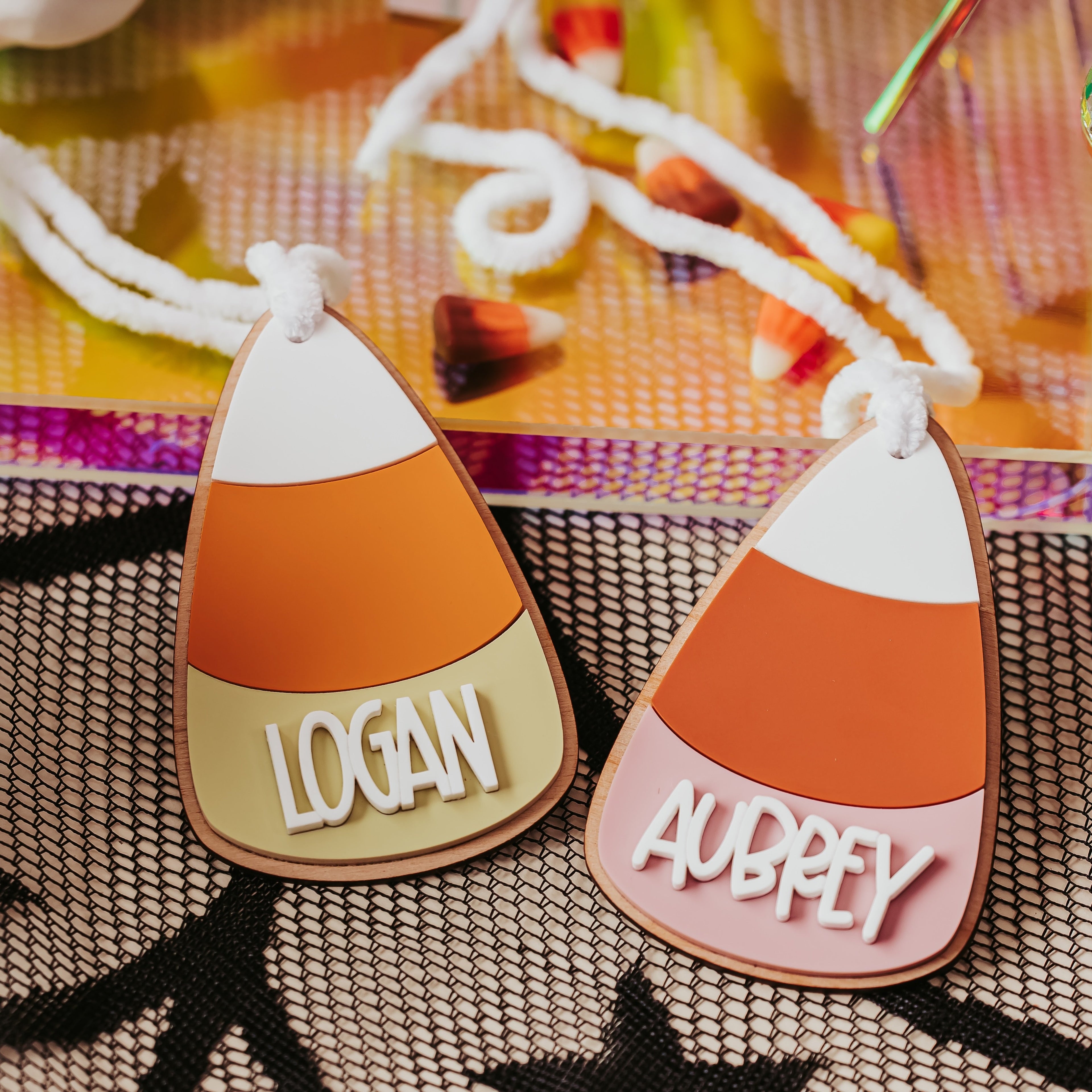Two personalized candy corn boo basket tags with the names 'LOGAN' and 'AUBREY' in white lettering, displayed on a black mesh tablecloth with Halloween-themed props.
