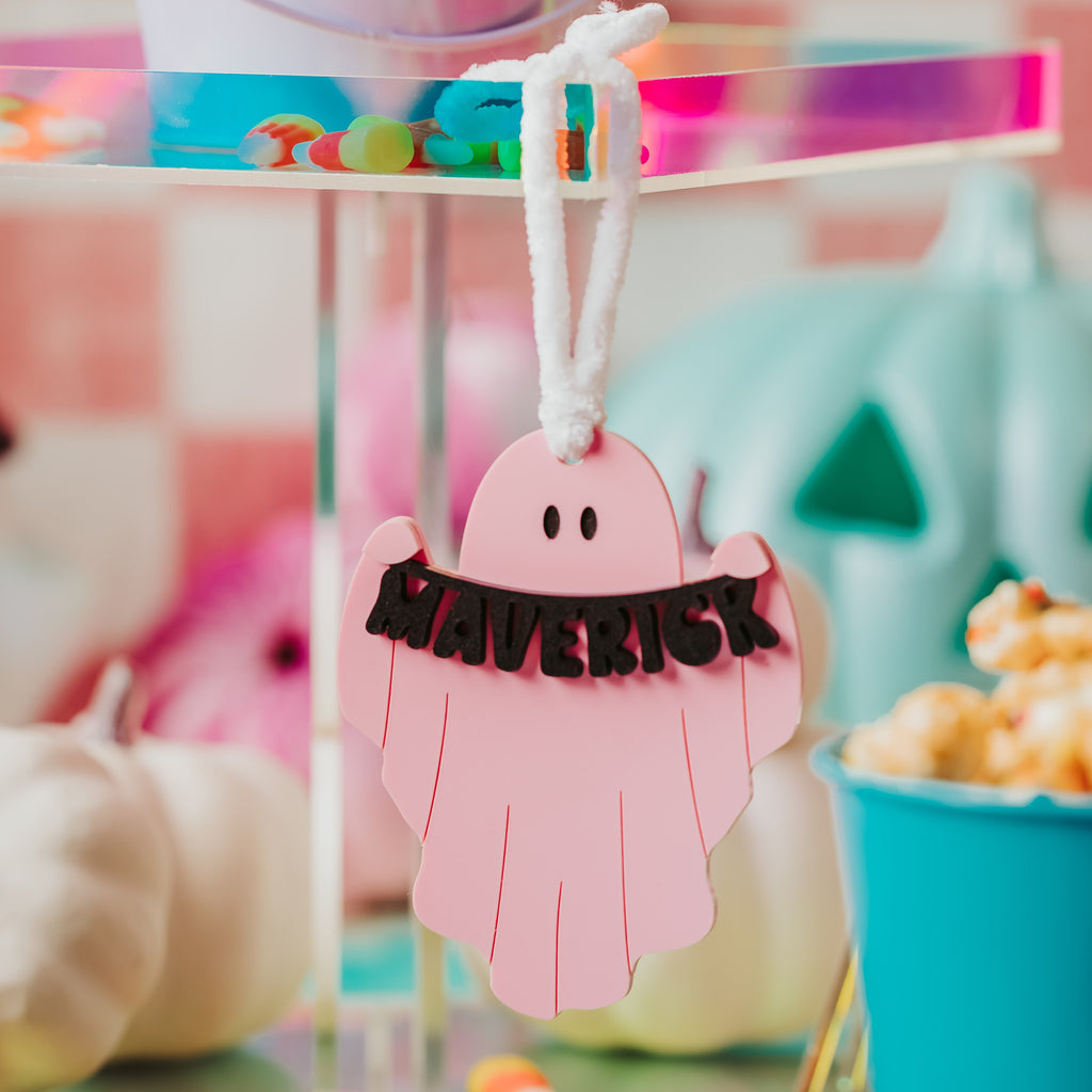 A pink ghost-shaped boo bag with the name "MAVERICK" in black letters, suspended by a white string on a clear acrylic stand in a colorful party setting.