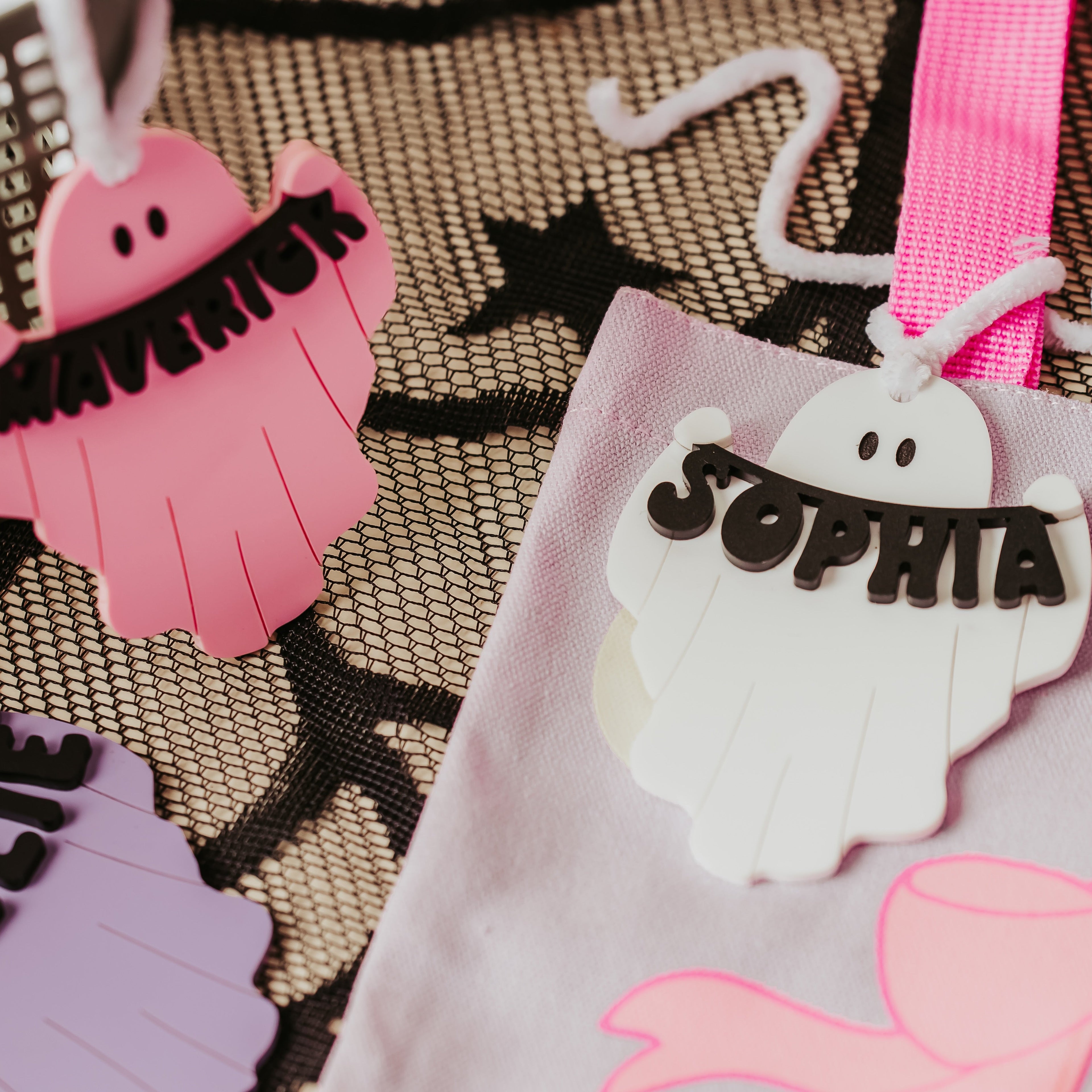 Close-up of a light pink fabric bag with a white ghost-shaped basket tag that says "SOPHIA" in black letters, alongside other ghost tags in pink and purple on a black-and-beige patterned surface.