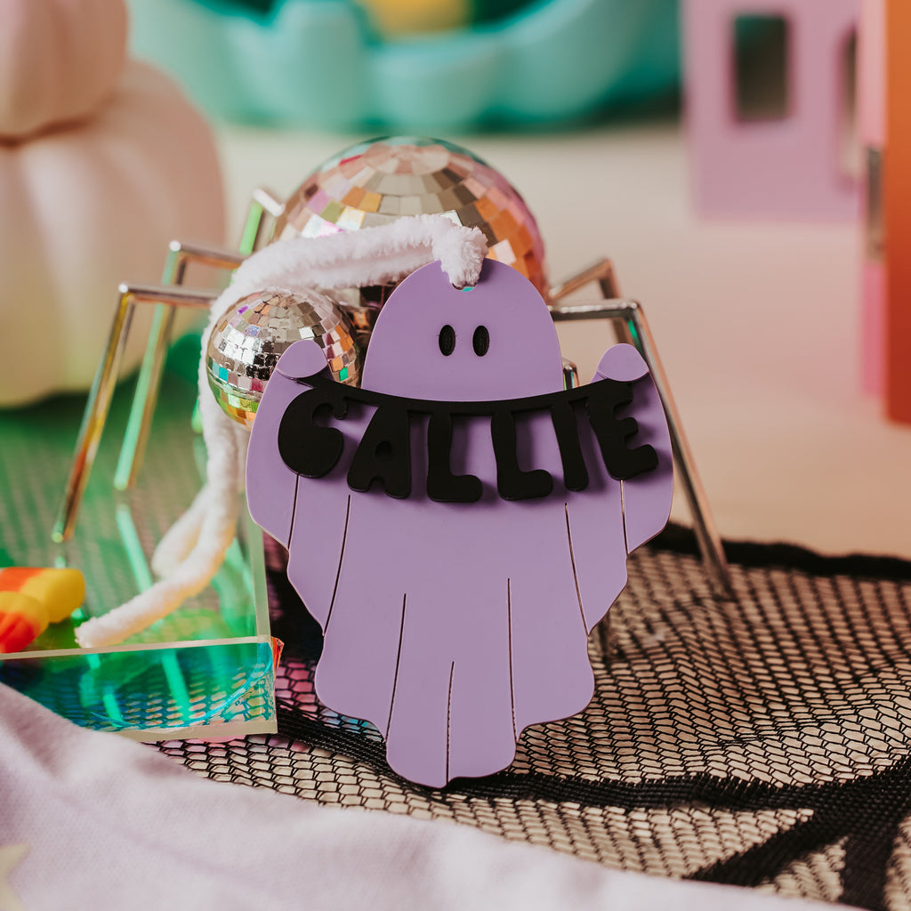 A Halloween-themed ghost boo basket tag with the name 'CALLIE' in black letters, displayed on a spider stand with a disco ball prop. The scene is decorated with a teal jack-o'-lantern and a pink spider cutout in the background.