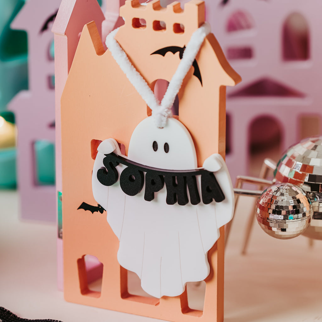 A Halloween-themed wooden stand featuring a peach-colored haunted house silhouette with a white ghost boo basket tag cutout in the center holding a black banner that reads 'SOPHIA'. The stand is styled with a black lace mat in the foreground and pastel-colored wooden house cutouts in the background.
