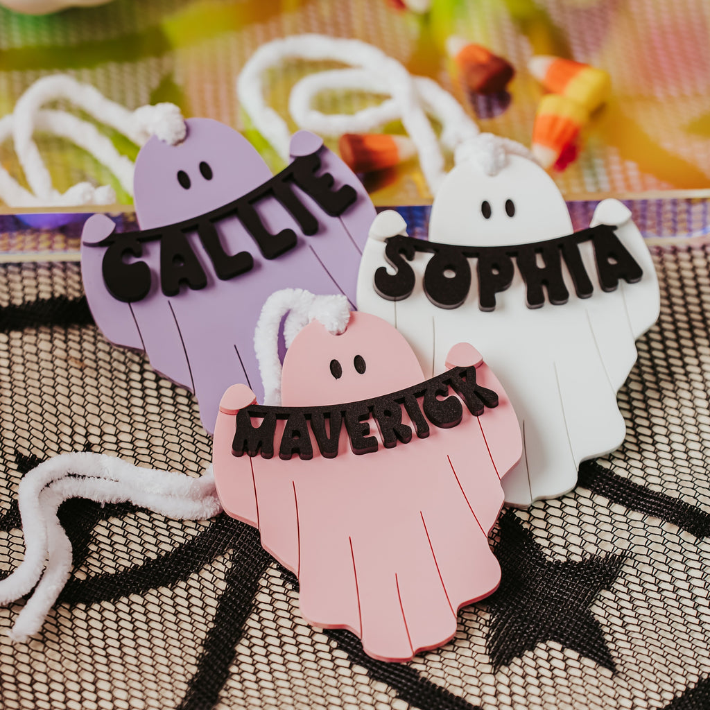 Three personalized ghost-shaped halloween basket tag in pastel purple, pink, and white, each with a black banner reading a name (Callie, Sophia, Maverick) and white string ties, displayed on a black spiderweb-patterned mat.