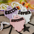 Three personalized ghost-shaped halloween basket tag in pastel purple, pink, and white, each with a black banner reading a name (Callie, Sophia, Maverick) and white string ties, displayed on a black spiderweb-patterned mat.