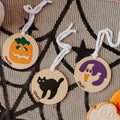 Three round acrylic Halloween sugar cookie bag tag with white hanging strings are displayed on a black spiderweb-patterned fabric. One ornament shows an orange jack-o'-lantern labeled 'Camryn', one shows a black cat labeled 'Tiffany"