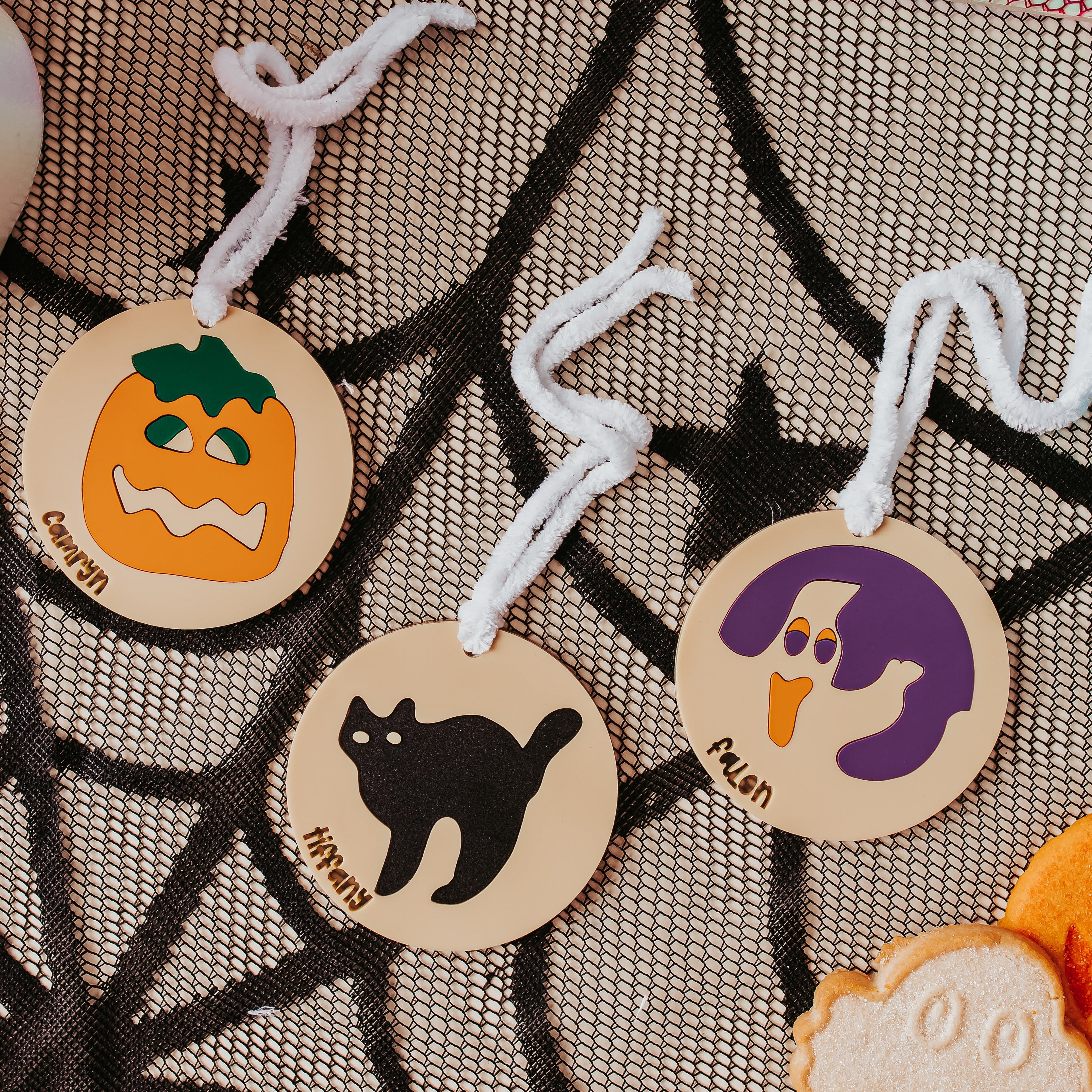 Three round acrylic Halloween sugar cookie bag tag with white hanging strings are displayed on a black spiderweb-patterned fabric. One ornament shows an orange jack-o'-lantern labeled 'Camryn', one shows a black cat labeled 'Tiffany"